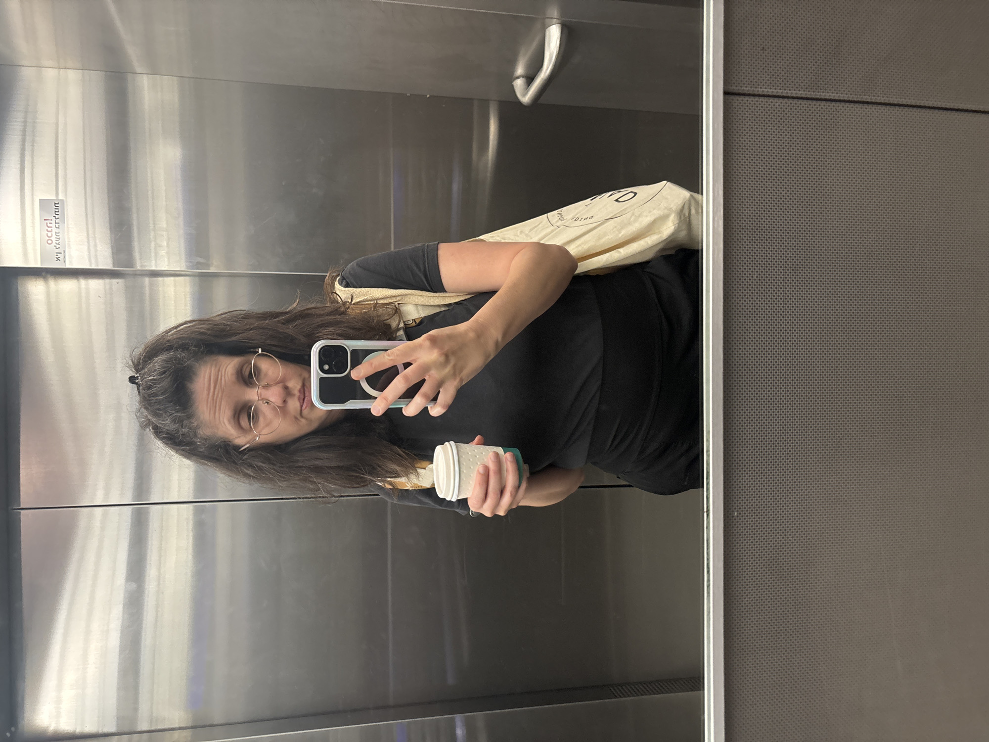 A woman in a black top and round glasses takes a selfie in a mirrored elevator in what is presumed to be Tel Aviv, Israel. She holds a white reusable coffee cup in her left hand and her phone in her right, partially obscuring her face. A beige tote bag is slung over her shoulder. The elevator walls are a mix of textured gray and reflective metal.