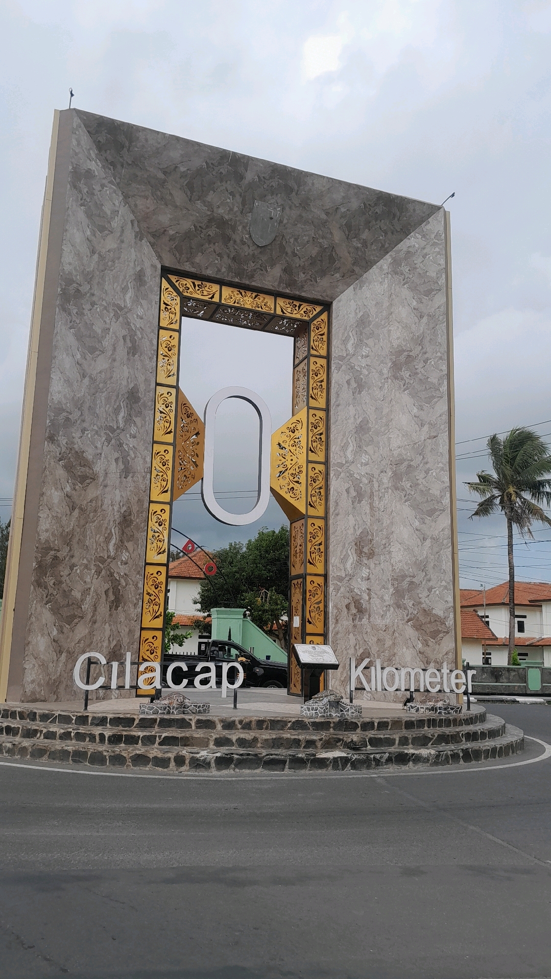 This is a medium shot, eye-level, outdoor photograph of the Zero Kilometer monument in Cilacap, Indonesia, under an overcast sky. The monument is a large, square structure with a marble-like facade and a large archway in the center. The archway is framed with gold ornate metalwork and contains a large white number "0." White text on the monument reads "Cilacap Kilometer." The monument is surrounded by a circular stone base, and a road curves around it. To the right of the monument, a tall palm tree sways in the wind. In the background, several buildings with orange tile roofs are visible, as is a black pickup truck. The overall scene conveys a sense of a public landmark in a coastal or tropical urban environment.