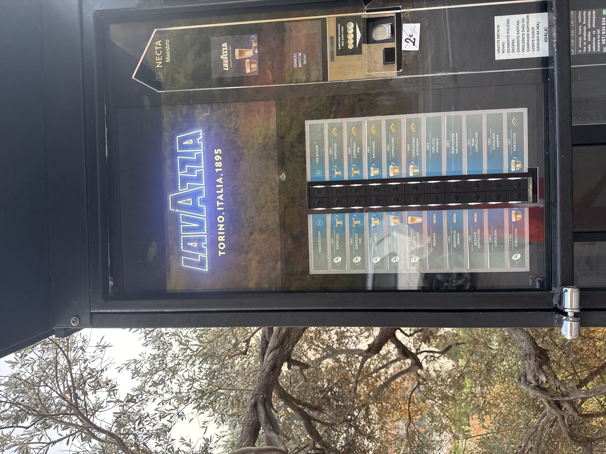 This is a medium shot, taken from a slightly elevated angle, of a vending machine. The machine dispenses coffee and other hot beverages, as indicated by the brand name "LAVAZZA TORINO, ITALIA, 1895" prominently displayed. The vending machine is black with a glass front, revealing the various beverage options and buttons. Several coffee types are listed with their prices in Euros, such as "ESPRESSO," "CAPPUCCINO," and "MOKACCINO." There's also an option for "IRISH COFFEE" and "ČOKOLADA" (chocolate). The machine accepts Euro coins, with a slot visible for insertion and a price of "2€" marked on a sticker. Instructions for using the machine are provided in Croatian.
Behind the vending machine, through the glass, a blurry image of coffee cups suggests the beverages being offered. To the left of the machine, the brand "NECTA Maestro" is also visible. The background outside the machine appears to be an outdoor setting with trees, possibly indicating the machine is located in a public space or cafe. The overall lighting suggests it might be daytime, although the exact time of day is not clear. The image is slightly tilted, suggesting it was taken handheld.
