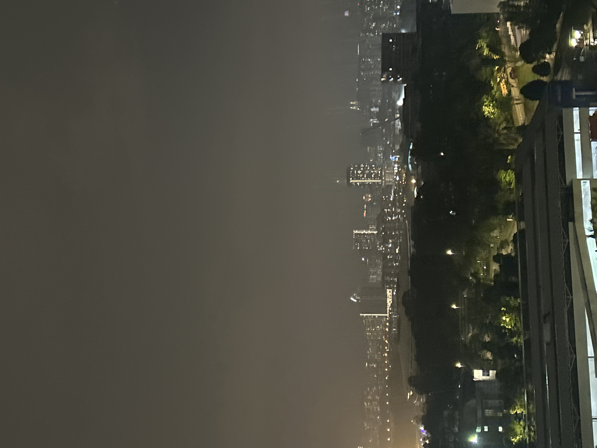 The image captures a nighttime cityscape of Kuala Lumpur, Malaysia, with the Petronas Twin Towers faintly visible in the distance, shrouded in haze. In the foreground, a dark expanse of trees obscures lower buildings, with scattered lights indicating streetlamps and structures. Further back, a dense cluster of city buildings with illuminated windows forms the urban skyline, punctuated by taller structures. The overall atmosphere is hazy and dark, typical of a humid night in a tropical city, with the city lights creating a diffused glow against the dusky sky.