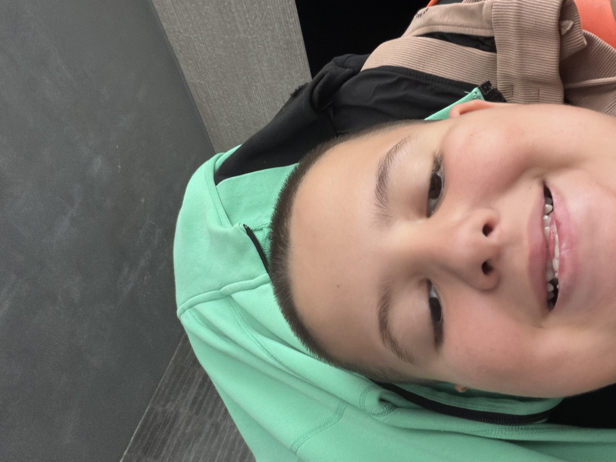 A close-up, slightly angled shot of a person's face. The person has short, dark hair and is wearing a light green hooded sweatshirt. They are smiling, revealing their teeth. Other clothing items with black and beige colors are partially visible in the background. The background also shows parts of a grey surface with a wooden grain pattern. The location in the city of Shumen, Bulgaria, is not discernible from the image.