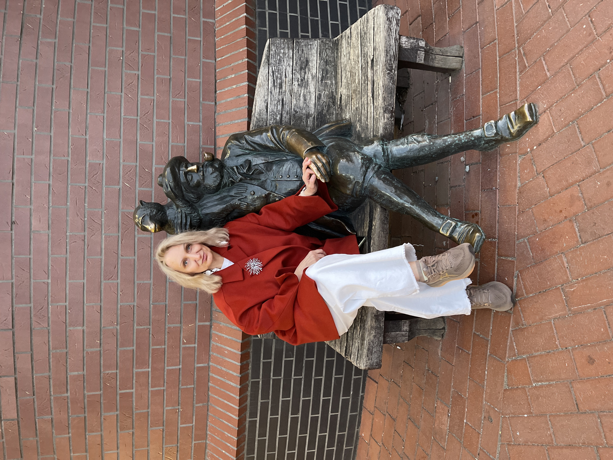 A woman sits on a wooden bench next to a bronze sculpture of a man lying down. The woman wears a red jacket, white pants, and tan sneakers. The sculpture depicts a man with a hat, lying on his back with one leg crossed over the other. The bench is made of weathered wood, and the background consists of brick walls. The location is Kaliningrad, Russia.