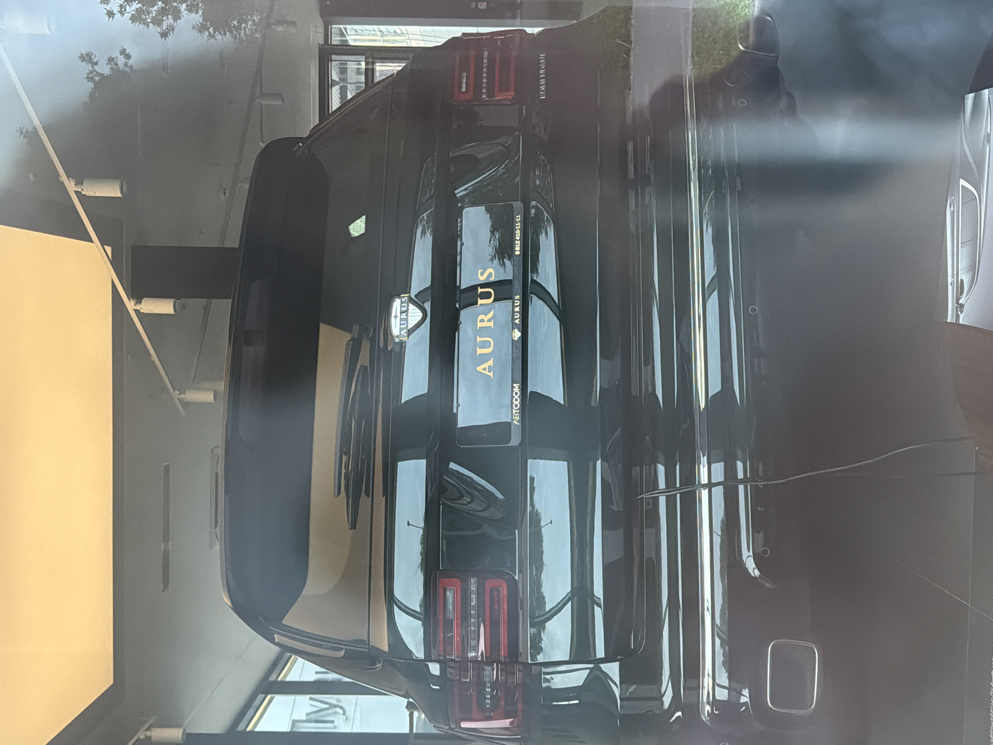 The rear view of a black Aurus luxury SUV is shown. The car has a gold license plate with the word "AURUS" in gold letters. The car has chrome accents and taillights that wrap around the sides. A reflective chrome exhaust pipe is visible on the lower left side of the bumper. The car is parked indoors, likely in a showroom, with bright overhead lighting and reflections of the surroundings on its polished surface.
