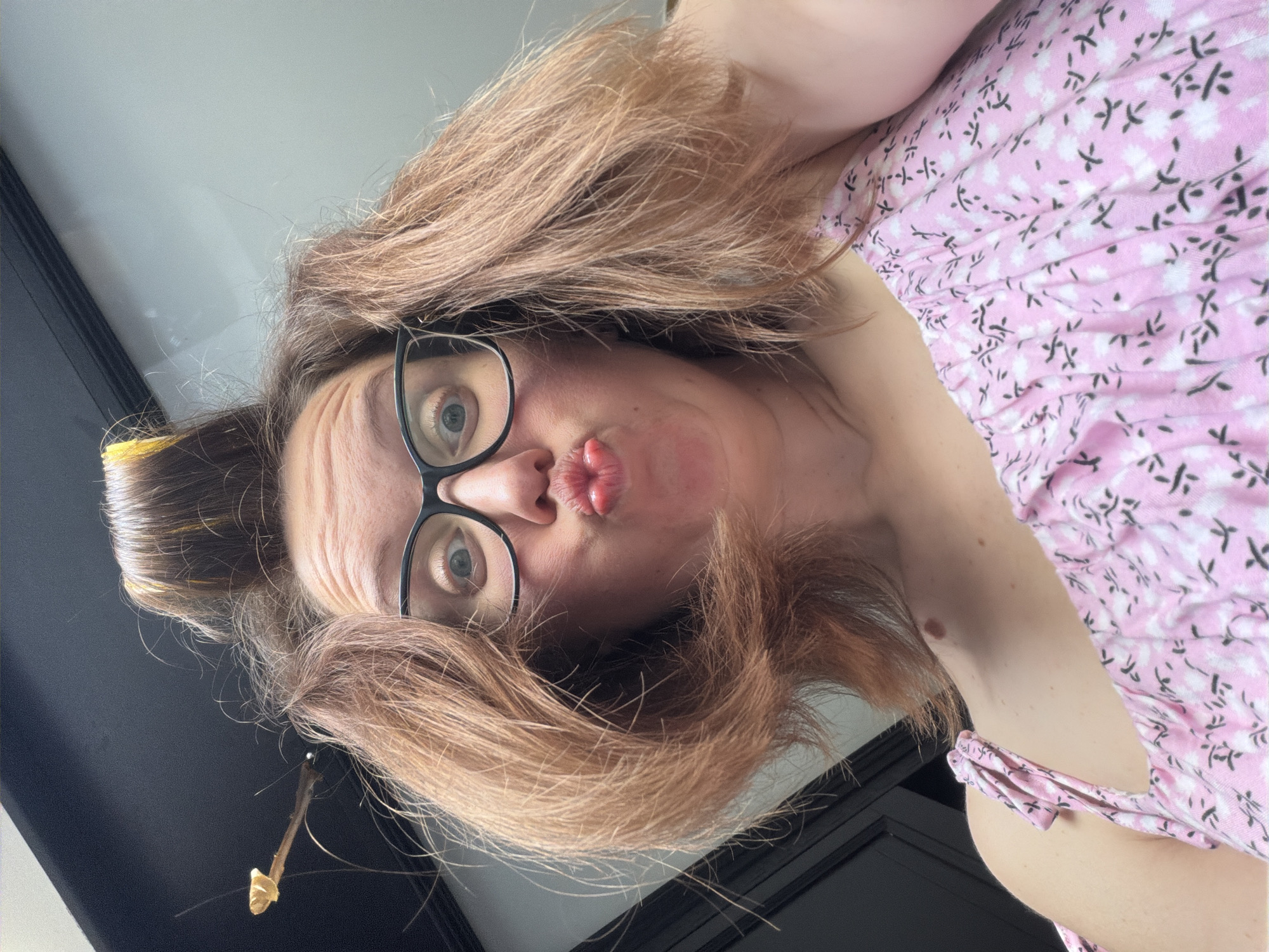 A low-angle, close-up, selfie-style shot shows a woman with light brown hair and glasses. Her hair is styled with rollers, and a small twig is stuck in her hair. She is wearing a pink floral top. She is puckering her lips. The background appears to be indoors, with dark walls or cabinets. The image does not contain any information about the city of Bila Tserkva, Ukraine.
