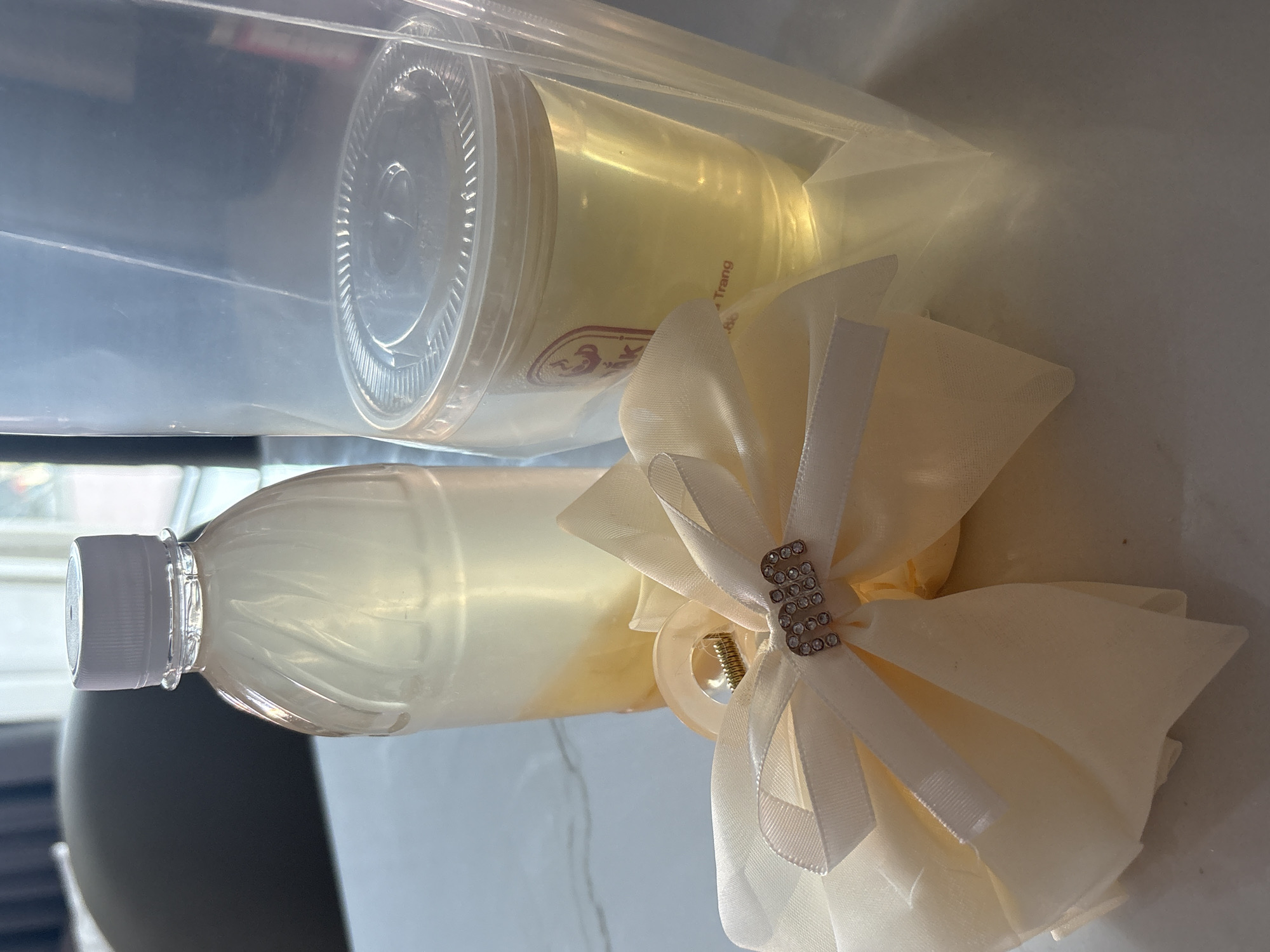 This still life image captures a collection of items placed on a light-colored, flat surface, likely a countertop, under bright, natural lighting.
The primary subjects include a clear plastic bottle with a white or grey screw cap, filled with a translucent, light yellow liquid. Adjacent to it is a clear plastic cup with a lid, also containing a similar light yellow liquid. This cup is enclosed within a clear plastic bag. On the side of the cup, a red logo featuring an animal or mythical creature is partially visible, along with some red text, including the word "Trang" and possibly a number "88". Given the provided context of "Bac Nha Trang Ward, Vietnam," this text likely refers to the location "Nha Trang."
Next to the beverages is a decorative, cream-colored fabric bow. Made of a sheer, soft material, it features a prominent, shimmering embellishment in the shape of the letter "M," adorned with small, clear rhinestones. A clear plastic claw clip mechanism is visible behind the bow, suggesting it is a hair accessory.
No specific activity or event is taking place; the items are simply displayed. The bright lighting suggests it is daytime. The background is softly out of focus, showing indistinct reflections and shapes typical of an indoor environment.