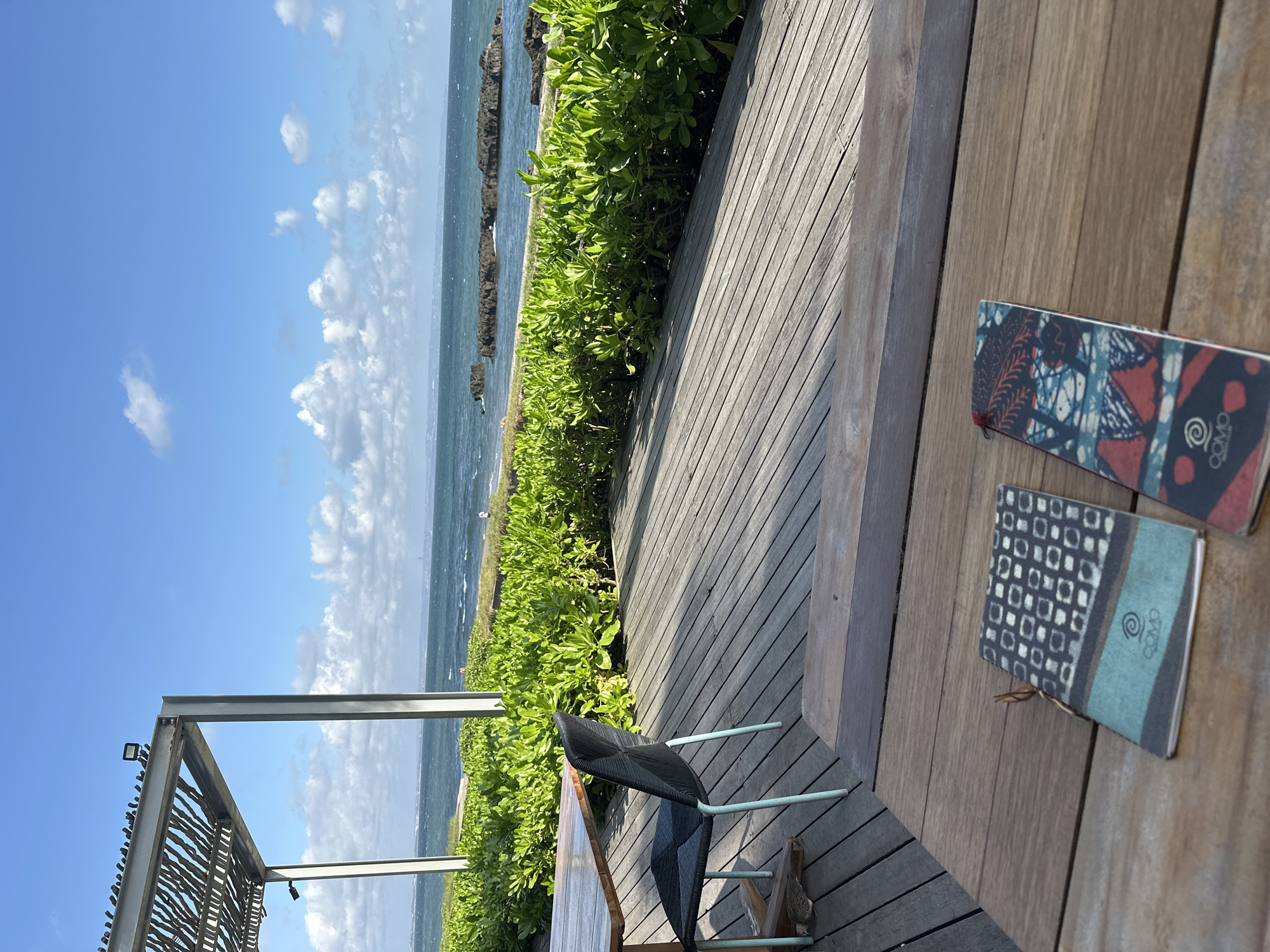 The image shows an outdoor scene, presumably in North Kuta, Indonesia, due to the presence of "COMO" branded notebooks. The view includes a coastal landscape with the ocean, vegetation, and a partially visible structure that resembles a pergola. A wooden deck with a chair occupies the foreground and right side of the image. Two small notebooks are placed on the table. The sky is blue with scattered clouds. No people are visible.