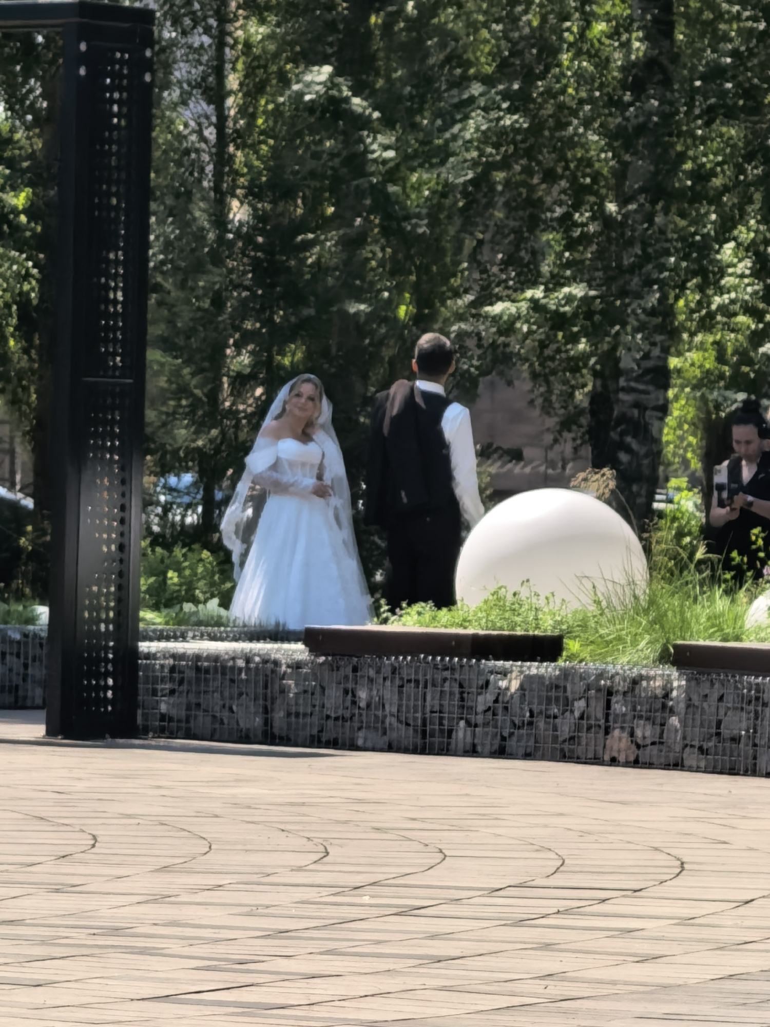 In Sterlitamak, Russia, a wedding party is captured in an outdoor setting. A bride, wearing a white wedding gown and veil, stands to the left, smiling at the camera. She is positioned in front of a gabion wall filled with stones. To her right, a groom in a white shirt and black vest is partially visible, facing away from the camera. Behind the couple, lush green trees form a backdrop, with a large, white spherical object visible in the mid-ground. To the far right, a person holding a smartphone is seen. The foreground consists of a paved area with a circular pattern.