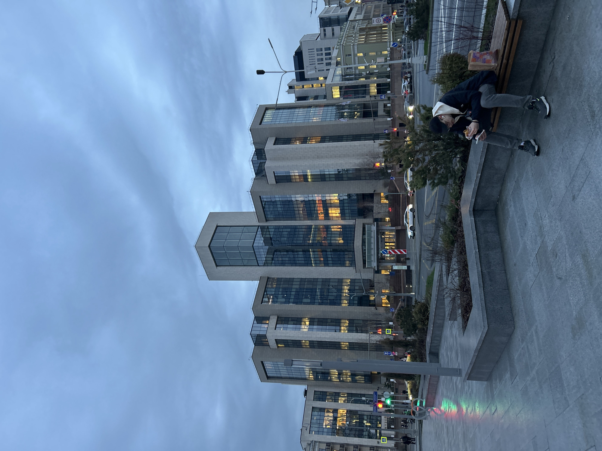 The image shows a street scene in Moscow, Russia at dusk. A person is sitting on a bench near a modern building. The building has a tiered structure with glass and stone facades. Lights are visible inside the building, suggesting that it is an office building. The person on the bench appears to be eating. Other buildings and street signs are visible in the background. The sky is overcast. A light post shows green, red and another color.