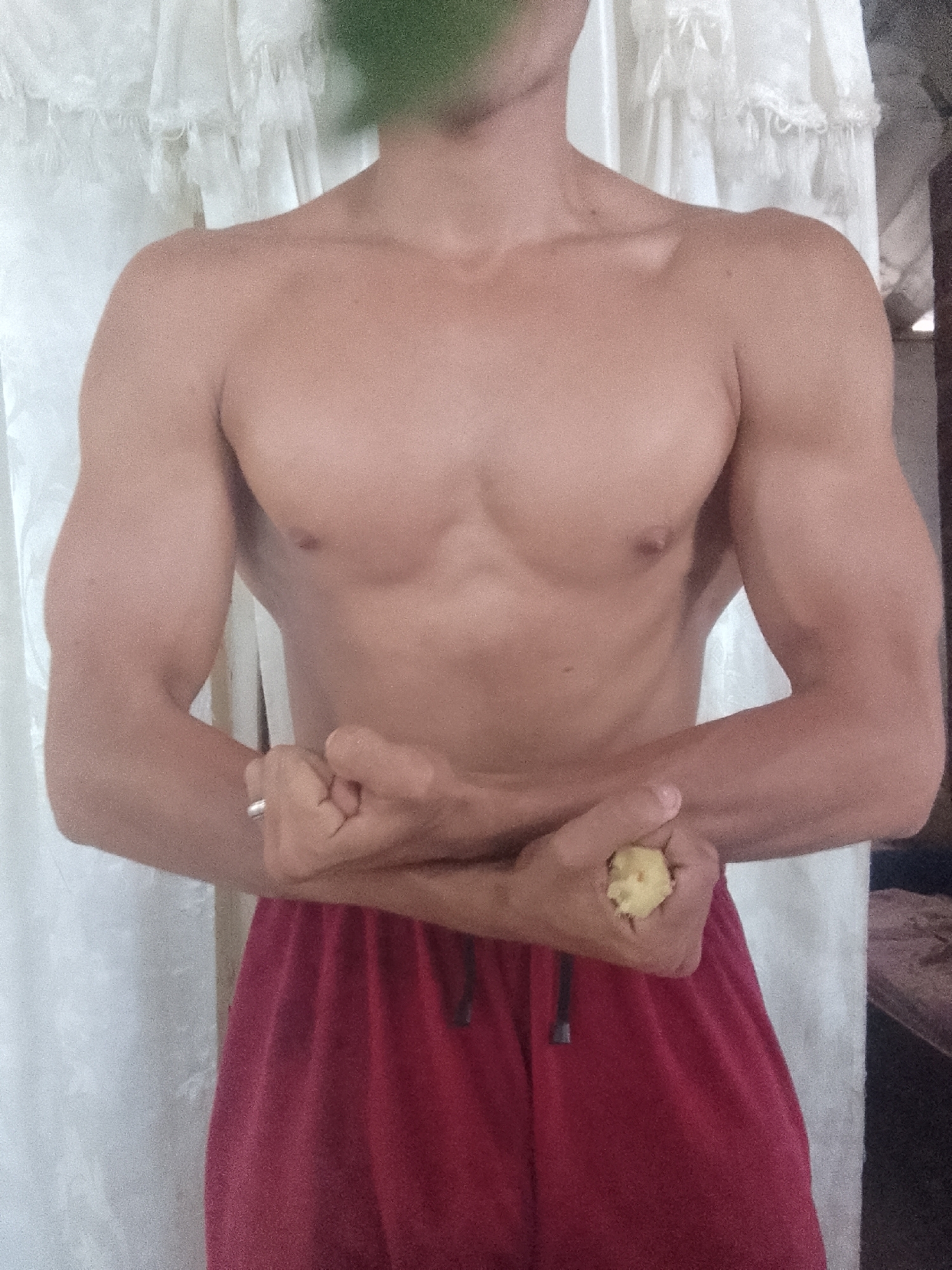 A shirtless man is standing indoors in front of a white curtain, wearing red shorts with a black drawstring. He is flexing his arms. He is holding a small, yellow object in his right hand. The top of his head is obscured by green pixels. The photo may have been taken in Juan Griego, Venezuela, but there is nothing in the image to confirm the location.