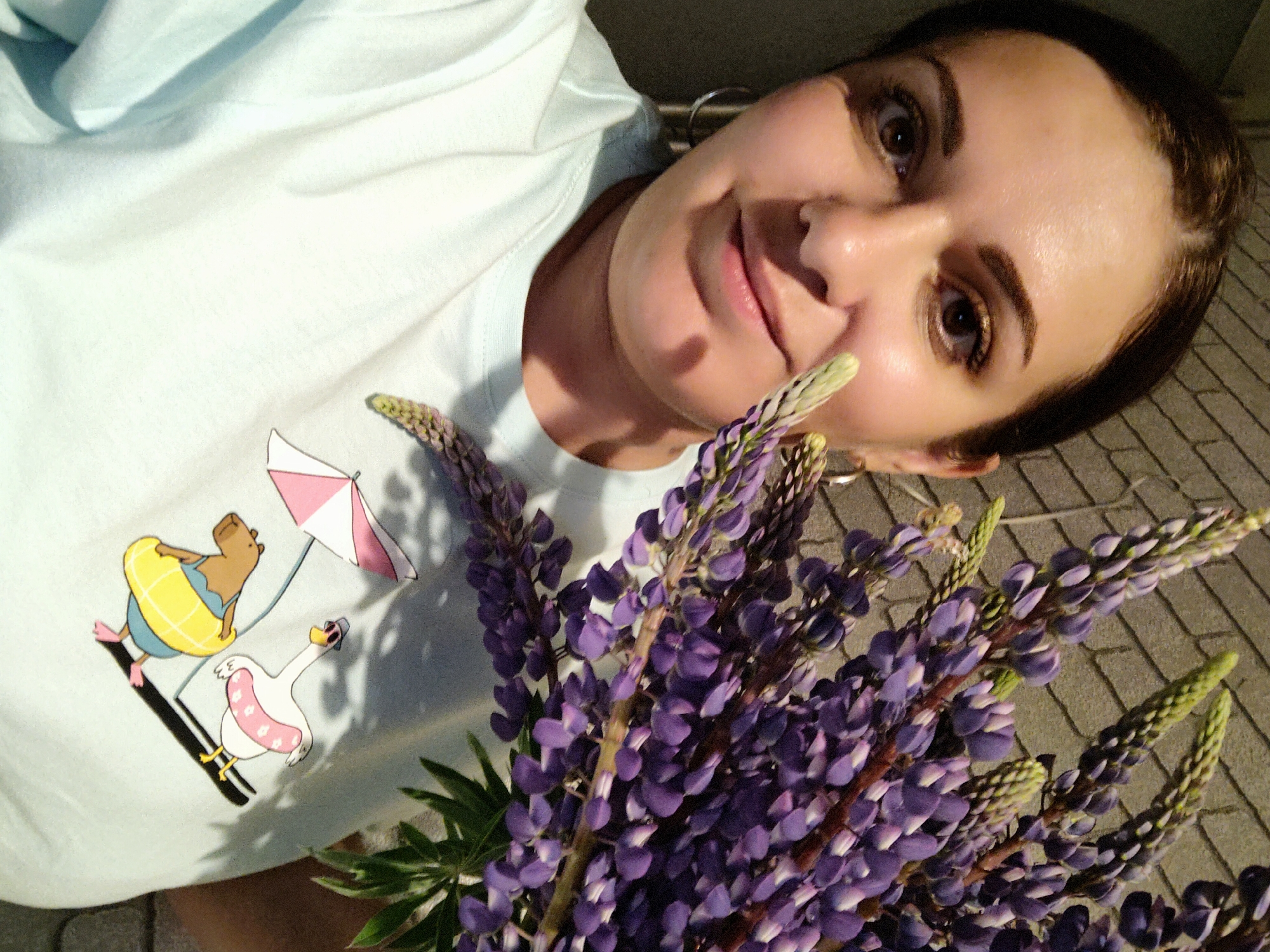 A woman is holding a large bouquet of purple lupine flowers. She is wearing a light blue t-shirt with a cartoon graphic of a capybara in a yellow inner tube and a swan with a pink inner tube floating on a black line, under a pink and white striped umbrella. She has brown hair pulled back, and brown eyes with gold eyeshadow. She is wearing silver hoop earrings. The background appears to be a paved area. The location is Марковский, Russia.