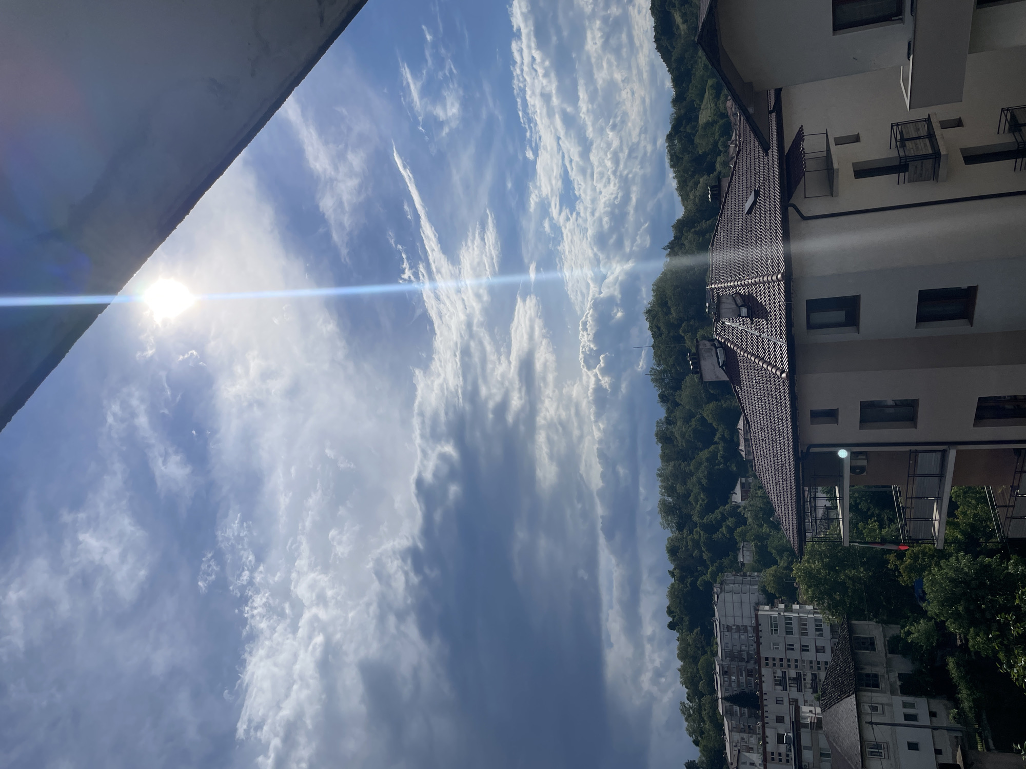 The image depicts a view in Zlatograd, Bulgaria. The sky is filled with scattered altocumulus clouds and a bright sun with a visible lens flare. Adjacent to the sky, there is a hillside covered in dense trees. On the right side of the image, there are several buildings, including residential apartments and houses, visible with their architectural details like roofs, windows, and balconies. The image is taken from a slightly tilted perspective, looking upwards and across towards the urban landscape and the natural environment. The photo is bright due to the daytime sunlight.