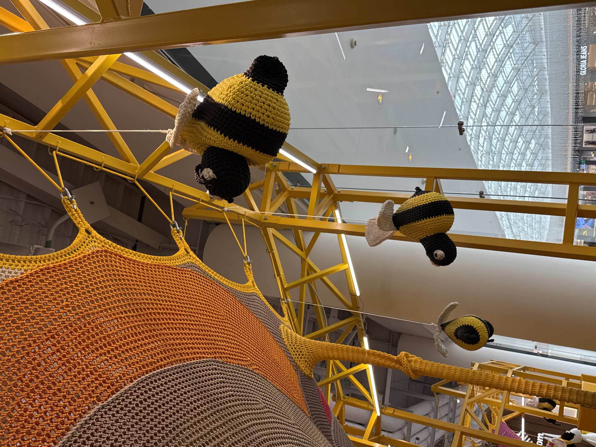 The image shows an indoor setting with a yellow metal structure and a large, multi-colored crocheted or knitted play area. Three crocheted or knitted bees are suspended from the structure. The play area has brown, orange, and yellow sections. The ceiling above is reflective, showing a large, multi-paned glass structure. The image was taken in the city of Kommunarka, Russia.