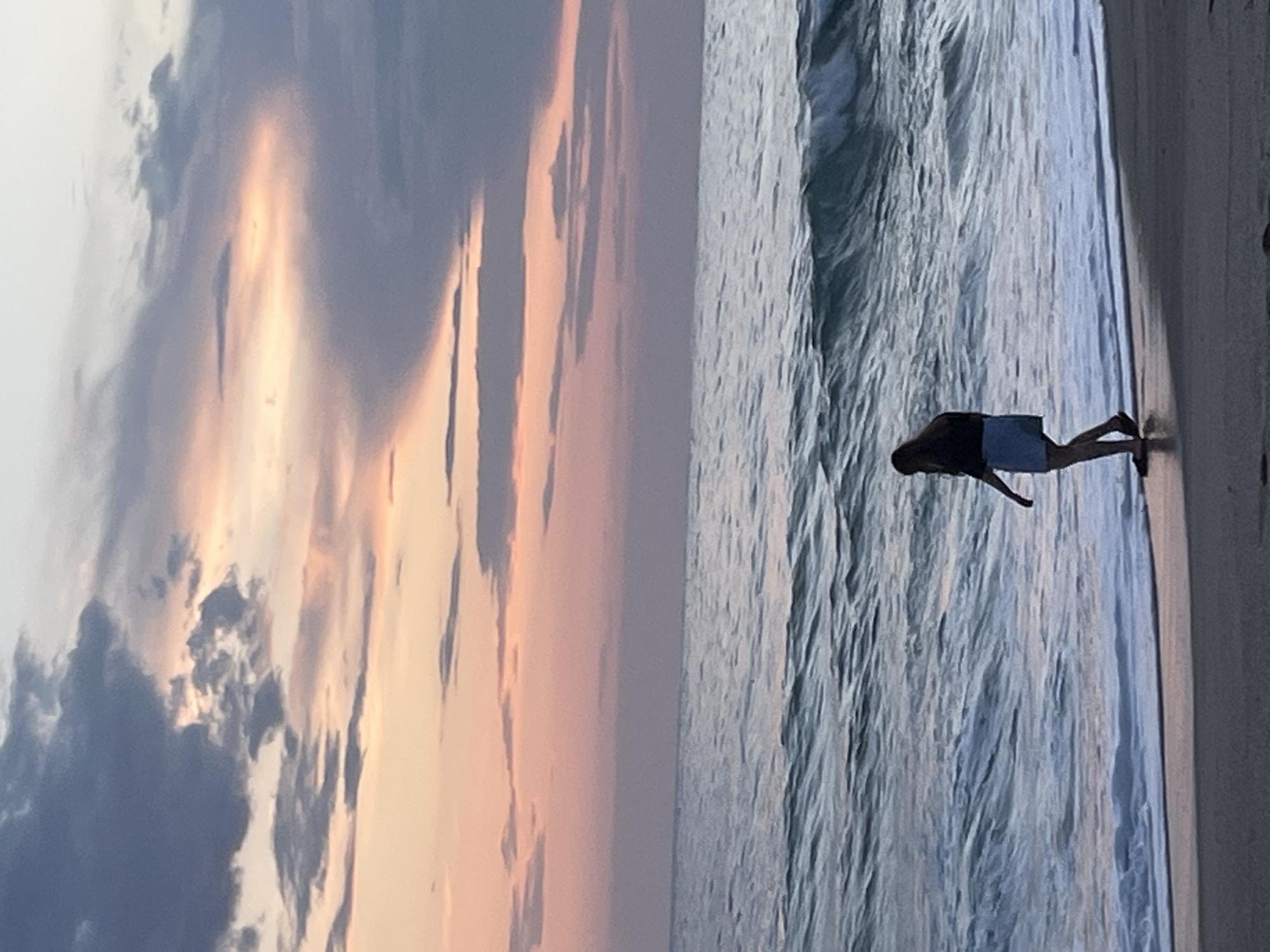 A photo shows a beach scene at dusk in Alanya, Turkey. To the left is a colorful sky with dark clouds and hints of orange and pink hues. To the right, a person is walking on the wet sand near the water. The person appears to be wearing dark clothing and light-colored shorts. The sea stretches out to the horizon with gentle waves. The photo is taken at an angle, rotated counterclockwise.