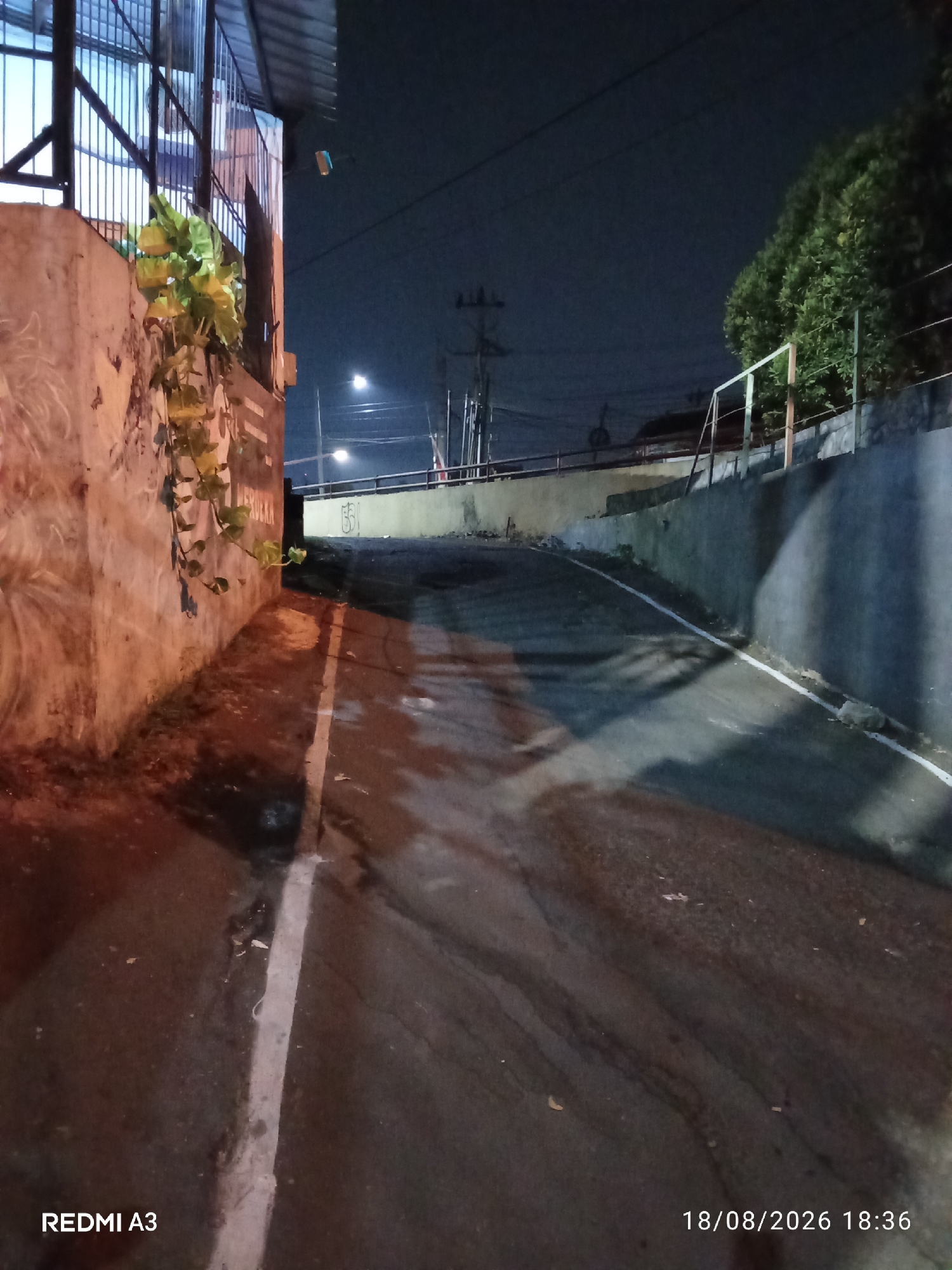 The image depicts a dimly lit, wet asphalt road at night, with a slight upward incline. On the left, a concrete wall with some graffiti and a climbing vine is visible. A metal railing runs along the top of the wall. To the right, another concrete wall lines the road, also with a railing. The sky is dark, indicating nighttime, and a few streetlights are present in the distance. The bottom left corner shows the text "REDMI A3", and the bottom right corner shows "18/08/2026 18:36". The scene suggests an urban environment, possibly a street in a residential or commercial area. Given the provided location "Purbayan, Indonesia", this is a nighttime street scene in that region.