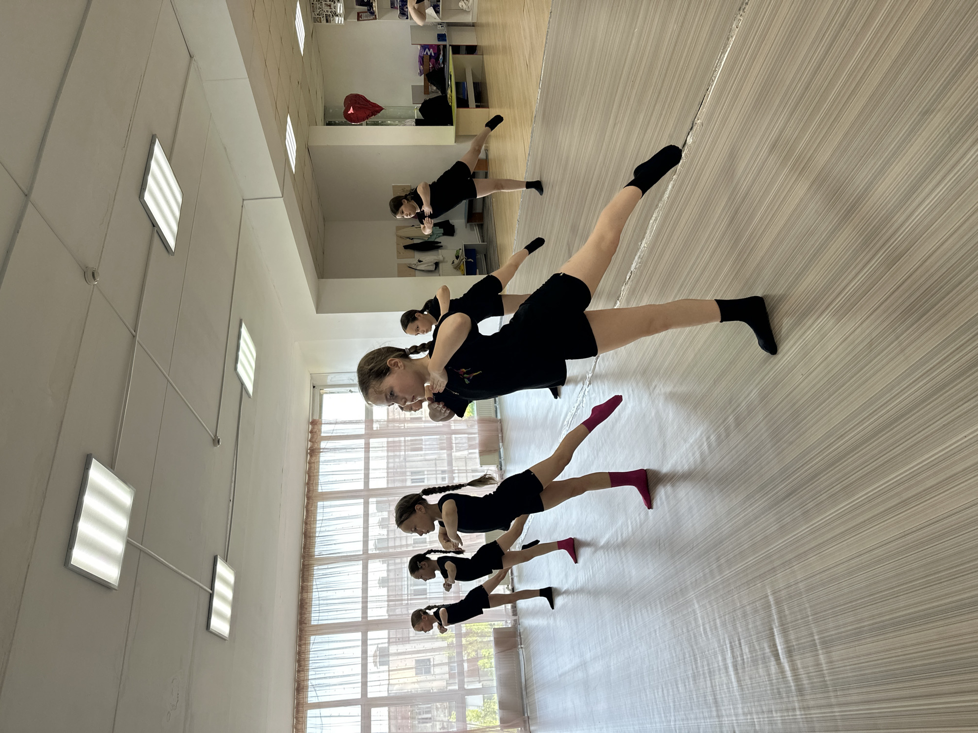 The image shows a dance class in Votkinsk, Russia. Six young girls are lined up in the middle of a dance studio with a mirrored wall. The girls are wearing black athletic wear and are in a plank position, with one leg extended and raised into the air. The studio is brightly lit by square panel lights and large windows.