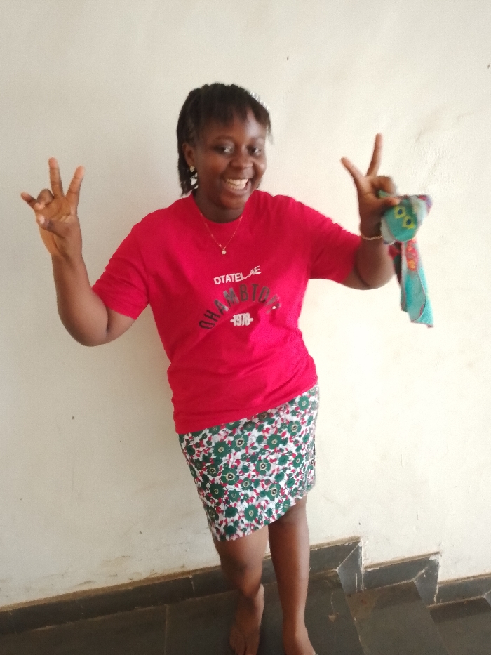 An adult female with a medium-dark skin tone and braided hair, styled with bangs, stands in an interior location within Umueshi, Nigeria. She is wearing a red short-sleeved t-shirt displaying text that includes "DTATELAE CHAMPION 1978", a gold-colored chain necklace with a small pendant, and a white, green, and red floral skirt that ends above the knees. She is barefoot. Both of her hands are raised and extended outwards, each displaying a two-finger 'peace' or 'victory' gesture. Her right hand (viewer's left) is empty, while her left hand (viewer's right) firmly grips a small, multi-colored fabric item. The individual has a wide smile with an open mouth. The background consists of a plain, light-colored wall, with a dark skirting board at the bottom that transitions into a visible set of dark-colored steps on the right side of the frame.
