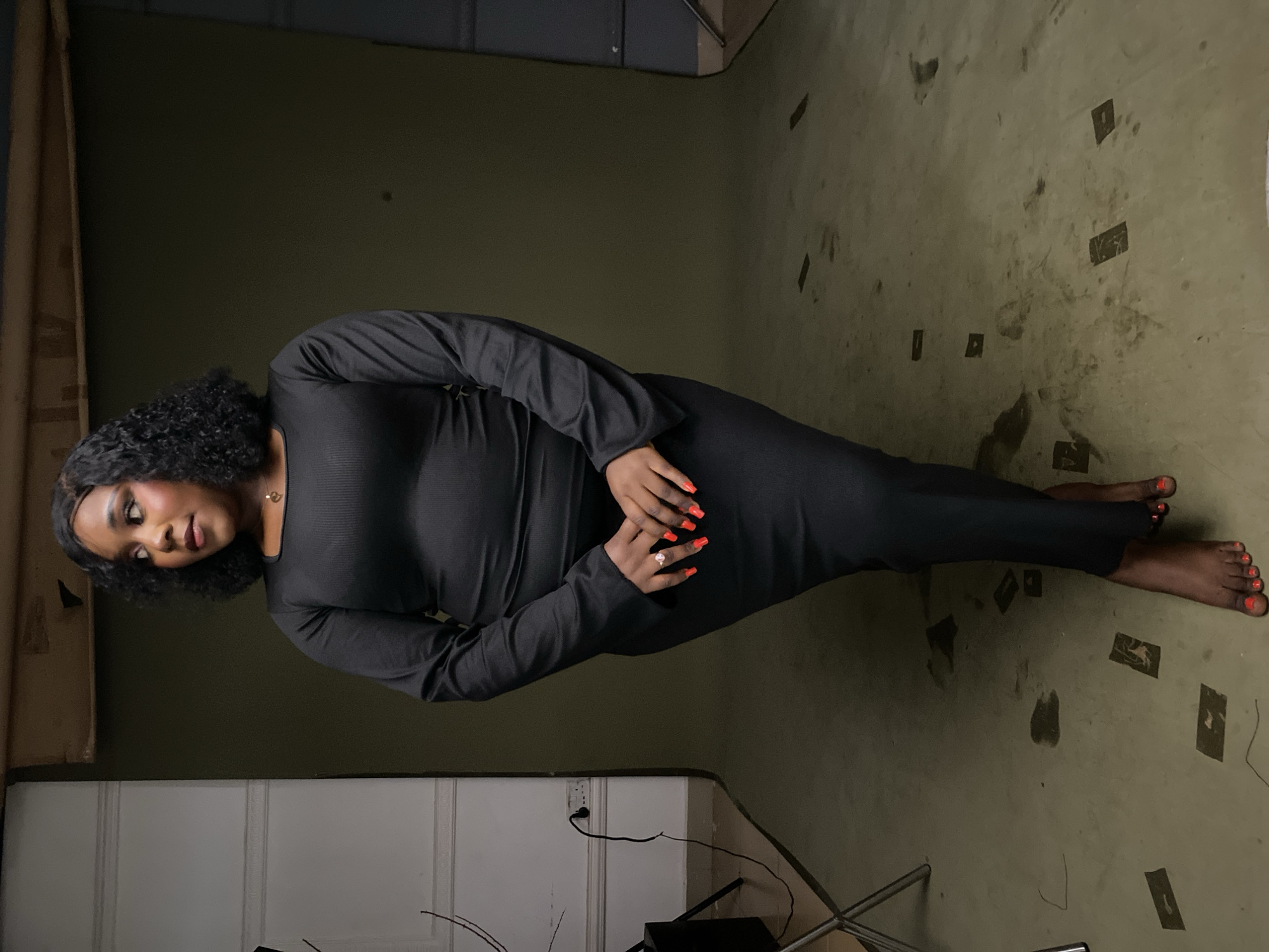 In a studio setting, a Black woman stands facing left, looking away from the camera. She is wearing a form-fitting, long-sleeved black dress that extends to her ankles. Her fingernails are painted bright red, and she wears a ring on her left hand. The background is a dark, neutral color, with a white wall on the left side featuring some decorative molding and electrical outlets. The floor is a light brown or tan color, with several dark rectangular shapes scattered across it. The lighting appears to be from studio lights, creating soft shadows. The overall atmosphere is that of a professional photoshoot.
