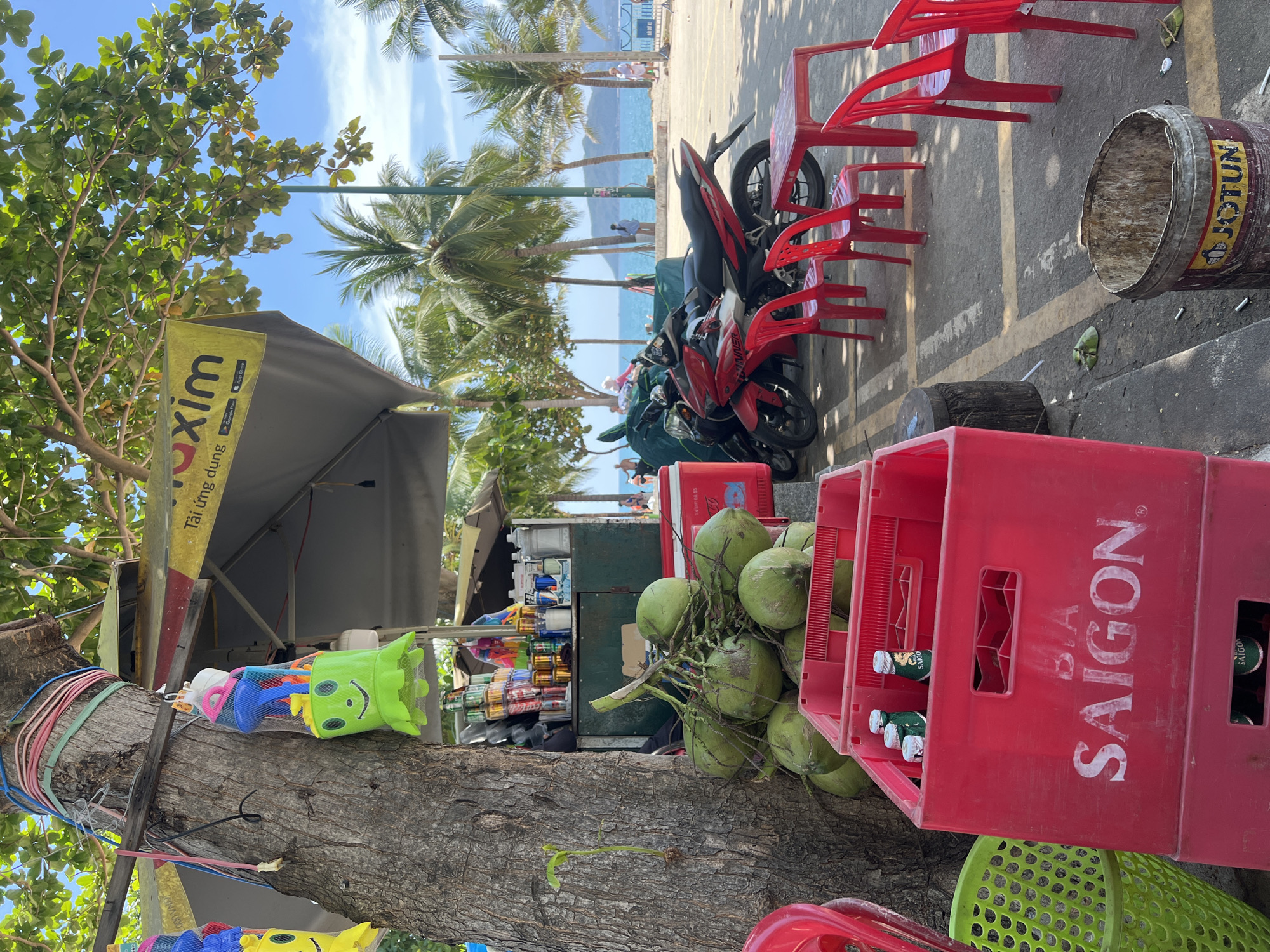 The image depicts a street scene in Nha Trang Ward, Vietnam. In the foreground, a tree trunk displays children's beach toys. Beside it is a vendor stand selling drinks and snacks. A pile of green coconuts rests near a red crate labeled "Saigon," containing beer bottles. A cooler is positioned behind the coconuts, and parked motorbikes are adjacent to red plastic chairs and tables. In the background, palm trees line a beach with people visible in the distance. The sky is blue and sunny.