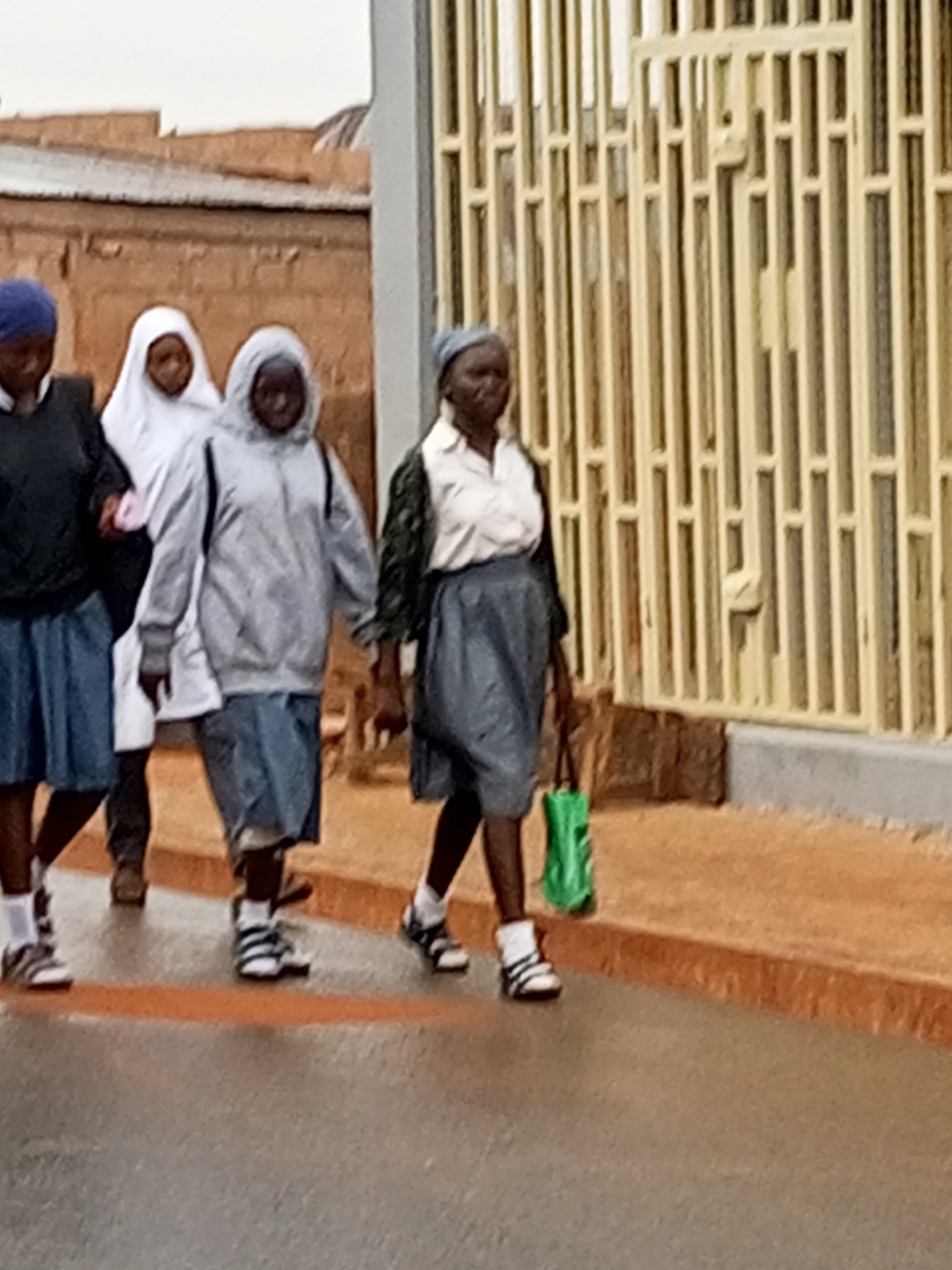 Four individuals, likely students, are walking along a wet paved surface next to a brick building and a yellow barred gate. The person on the far left wears a blue head covering, a dark jacket, and a blue skirt. Next to them is a person in a white hijab and a white robe. The third person from the left wears a grey hooded jacket and a blue skirt, and the person on the far right wears a white shirt, a dark patterned jacket, and a blue skirt, carrying a green bag in their right hand. All appear to be moving in the same direction. The ground is reflective, indicating recent rain. The background includes a rough-textured brown brick wall and a structure with vertical yellow bars.