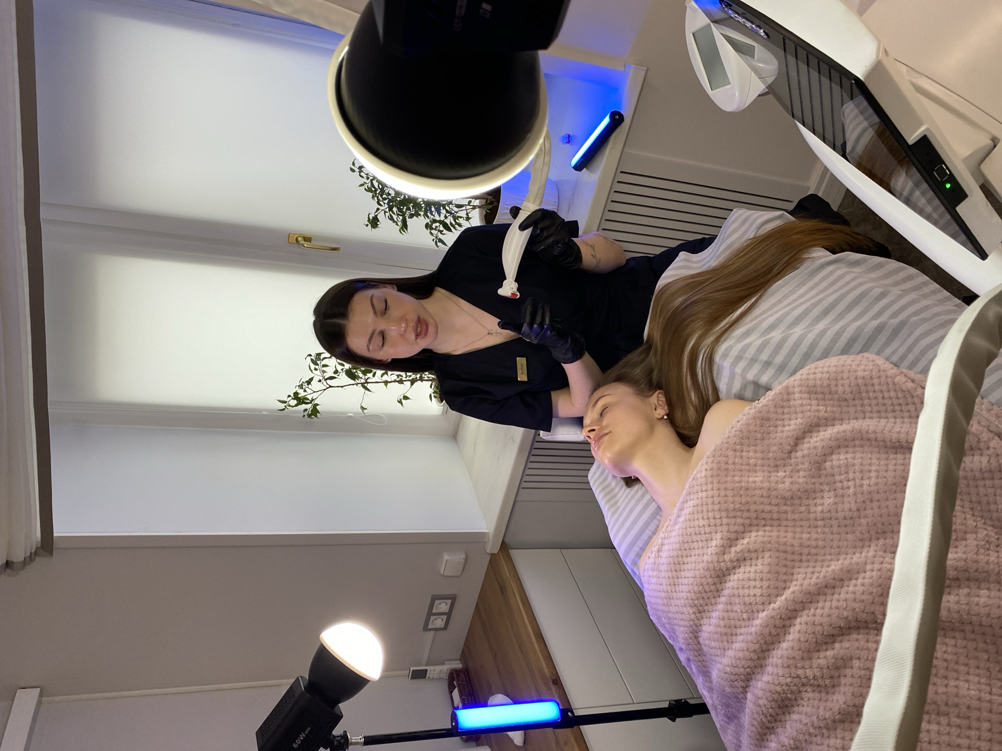 The image depicts a professional beauty salon or clinic setting, with two women present. A female practitioner, identified by a nametag reading "Anissa," stands dressed in a black uniform and black gloves. She has dark hair and is focused on a client, holding a white device with a red tip.
The client, with long brown/blonde hair, is lying on a treatment bed covered with striped bedding and a pink, textured blanket. Her eyes are closed, suggesting she is relaxed or receiving a treatment.
The room is well-lit by several artificial light sources, including a large studio light with a reflector positioned near the practitioner, a smaller spherical white light on a stand labeled "60W," and two blue LED tube lights on stands. Specialized equipment is visible, including a large white and black machine with a screen and grilles next to the client's bed. In the background, there's a window with a white frame and a gold handle, along with a potted plant. The setting appears modern, clean, and professional, designed for cosmetic or wellness services.