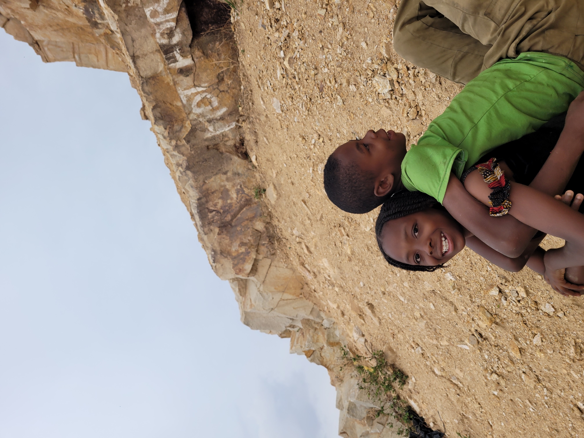 Two children are hugging on a rocky, sandy terrain in Barakin Akawo, Nigeria. A rock cliff is visible on the left. One child wears a green shirt and the other has braided hair and a colorful beaded bracelet. An adult's legs are partially visible above the children.