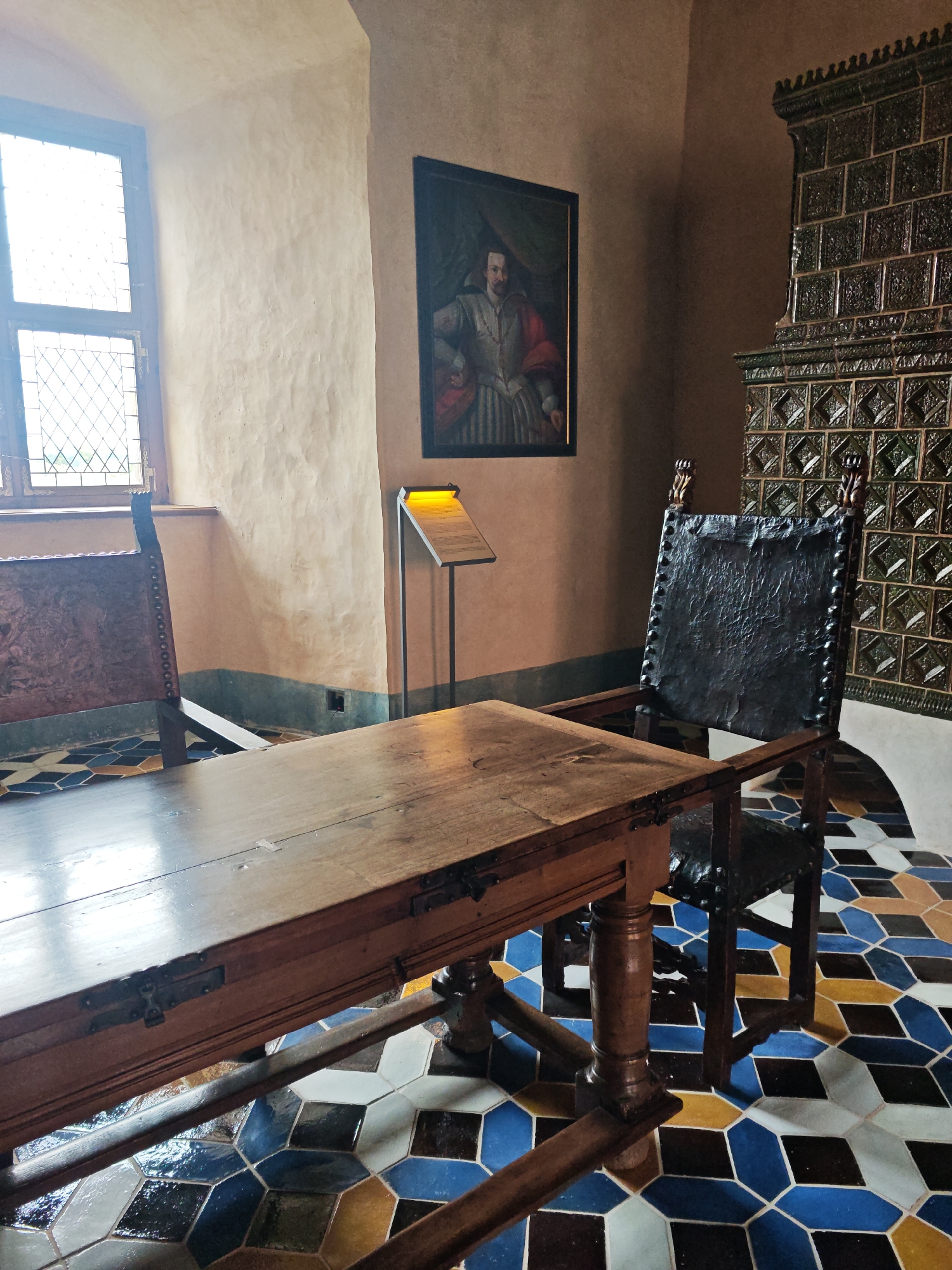 This image captures a room within Bauska Castle, Latvia, furnished with historical objects. In the foreground, a large, dark wooden table with metal latches sits on a floor adorned with a geometric pattern of colorful tiles in shades of blue, white, yellow, and brown. To the right of the table is a tall, dark leather chair with a high back, studded with metal rivets, and carved wooden armrests. To the left of the table, partially visible, is another wooden chair with a decorative back.
Dominating the background wall is a large, dark green tiled stove. Above the stove and slightly to the left hangs a framed portrait of a man in historical attire, featuring a red, white, and gold costume. To the right of the portrait is a simple, illuminated sign on a stand, likely providing information about the exhibit or the room. A tall window with diamond-shaped panes allows natural light to enter the room, illuminating the textured, light-colored walls. The overall impression is of a well-preserved historical interior.