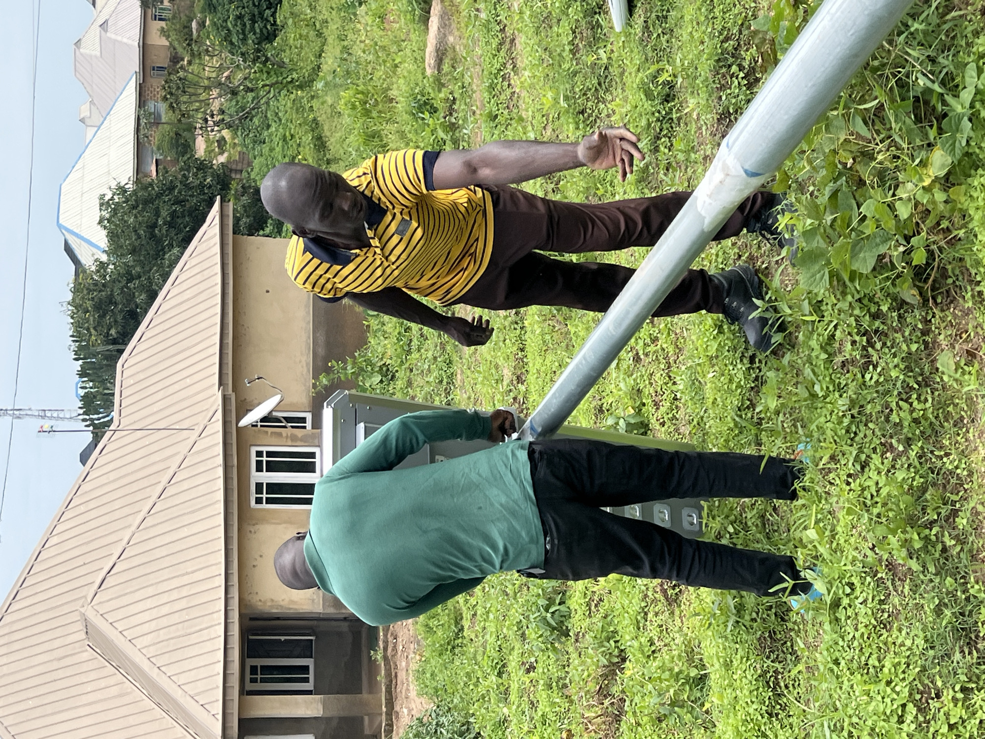 Two Black men are standing in a grassy area near a building with a light brown, corrugated metal roof. One man, on the left, wears a green long-sleeved shirt and black pants. He is facing away from the camera, with his hands on a silver-colored rectangular object. The second man, on the right, wears a yellow and black striped polo shirt and brown pants. He is facing the first man and appears to be gesturing towards a large, silver-colored pipe that runs diagonally across the foreground. The man on the right is wearing black boots. The environment appears to be outdoors, with lush green vegetation and other buildings visible in the background. The scene is likely in a residential area of Dong, Nigeria.