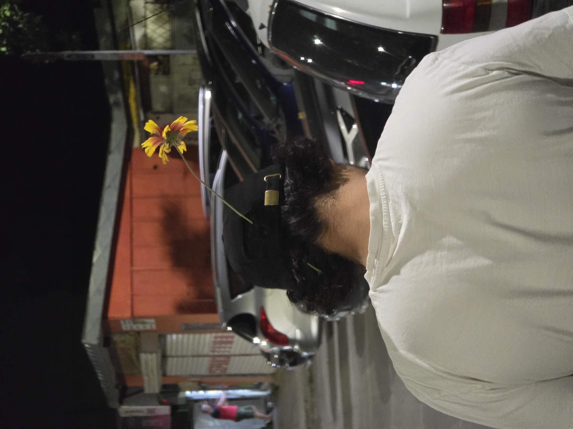 The image, taken at night in Varna, Bulgaria, shows the back of a person wearing a light-colored t-shirt and a black baseball cap. A yellow and red flower stem is stuck in the back of the cap, with the flower itself extending upwards. The person has curly dark hair visible around the cap. In the background, several cars are parked in what appears to be a street or parking lot. A red and orange building with closed shutters is visible on the left side of the frame, and a person in a red shirt and shorts can be seen near a doorway. The lighting suggests an urban environment at night.
