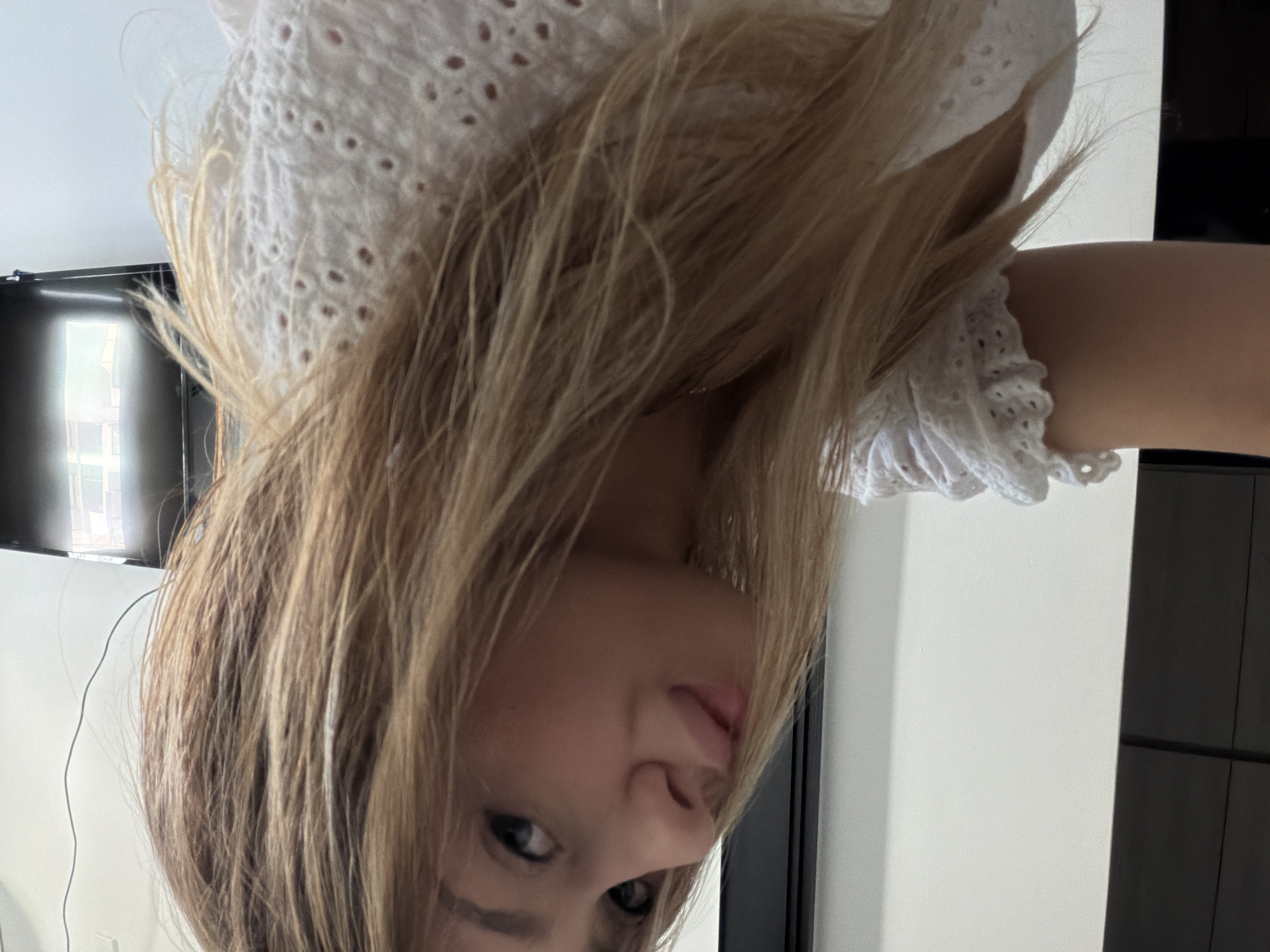 An over-the-shoulder selfie shows a young woman with blonde hair, looking towards the camera with her head tilted. She is wearing a white, eyelet-detailed top. The background is a plain white wall, with a dark, flat-screen television on the right. The lighting suggests an indoor setting, possibly an apartment. No specific location in Da Nang, Vietnam, is discernible from the image.