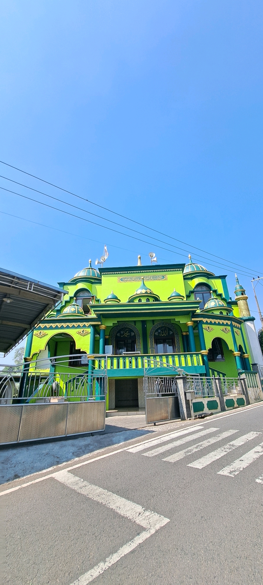 A vibrant green mosque with green and gold accents stands under a clear blue sky. The building features multiple domes and minarets, characteristic of Islamic architecture. In front of the mosque, a pedestrian crossing with white stripes marks the asphalt road. A metal fence separates the mosque's entrance from the street. Electrical wires stretch across the upper portion of the image, and a small canopy structure is visible on the left side. The mosque is located in Watuagung, Indonesia.