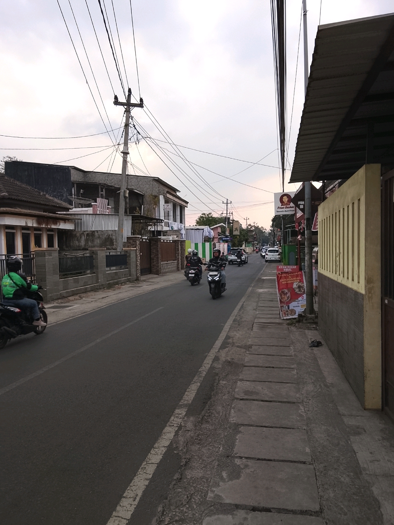 The image shows a street scene in Salatiga, Indonesia, under a cloudy sky. A paved road runs through the center of the frame, with a sidewalk on the right. Several motorcycles are visible, with riders on them. One motorcycle is in the foreground on the left, with a rider wearing a green jacket. Another motorcycle is further down the road on the left, and two more are in the distance on the right. Buildings line both sides of the street. On the left, there are houses with fences. On the right, there is a building with a corrugated metal roof and a yellow facade, featuring signage for "Bebek Fried Chicken" and "Tahu campur". There are utility poles with numerous wires overhead. The overall impression is a busy urban street with moderate traffic and pedestrian activity.