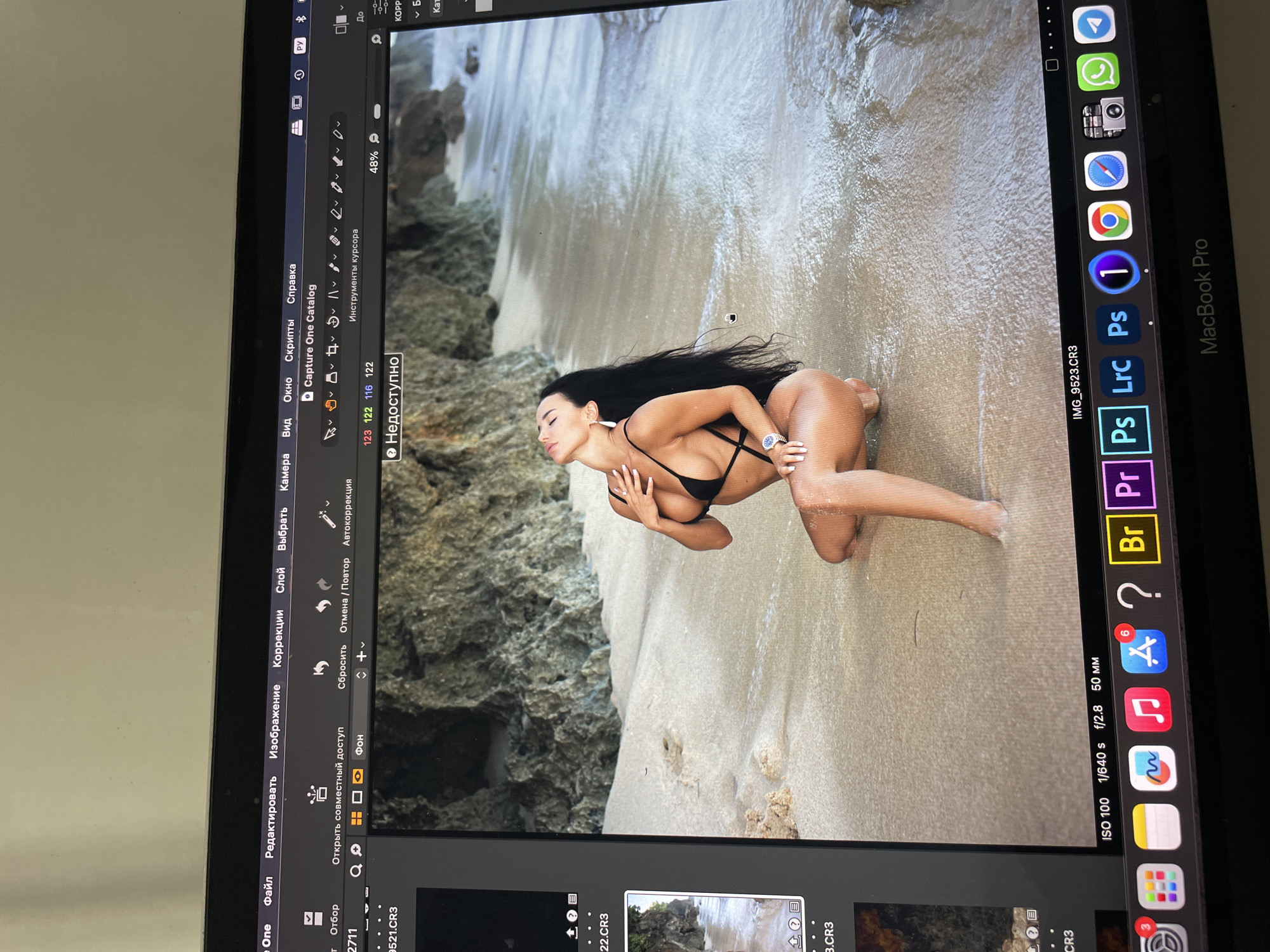 A woman in a black bikini is lying on a sandy beach, partially in the water, in front of some rocks. She has long black hair and is looking to the left. A Macbook Pro is open, displaying the image in Capture One software. The Macbook Pro is on a white surface. The operating system language of Capture One is set to Russian.