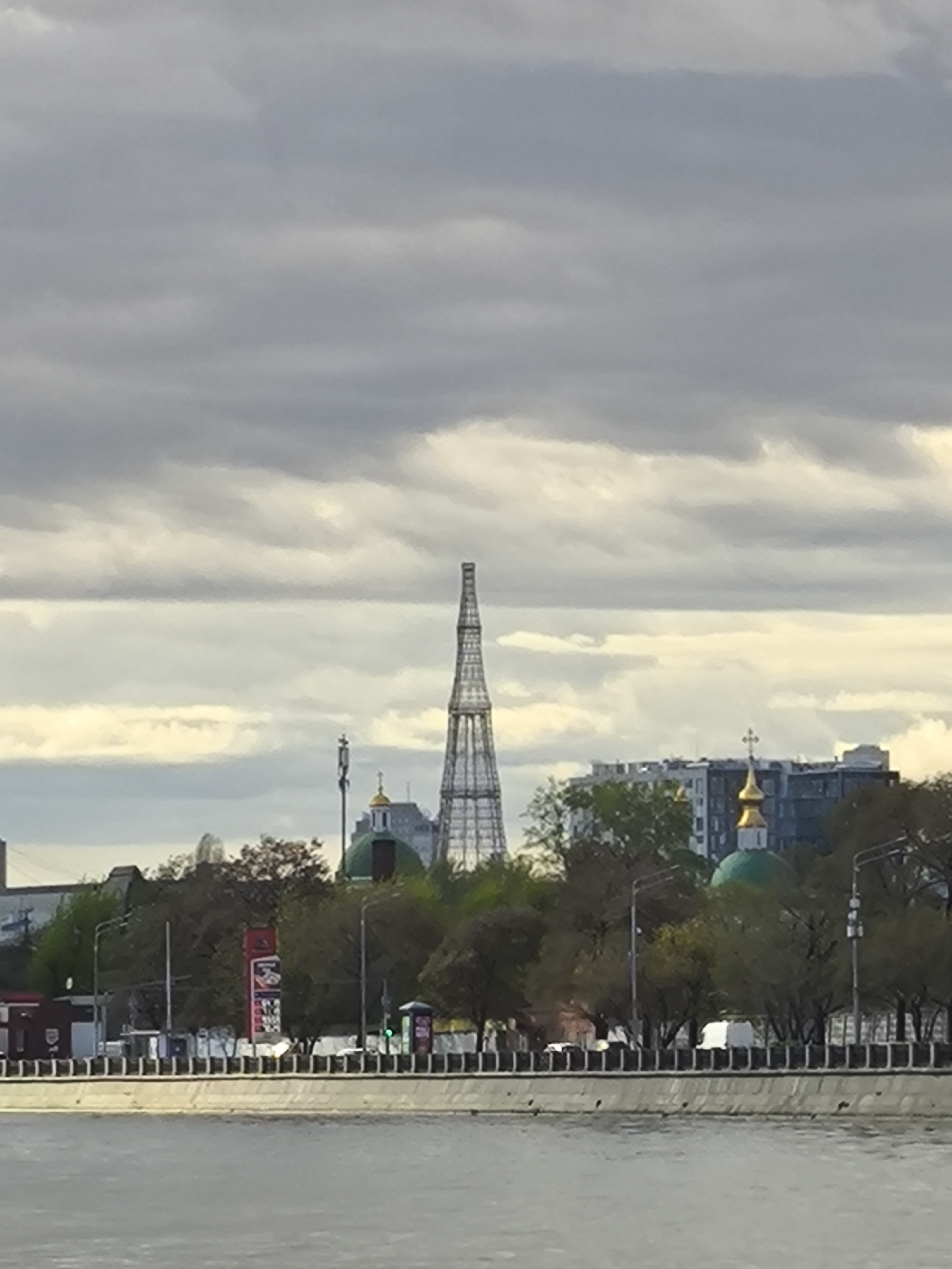The image depicts a cityscape in Moscow, Russia, with a river in the foreground. Dominating the skyline is the Shukhov Tower, a lattice steel structure. To its right are visible the green domes of a Russian Orthodox church. Buildings are nestled among trees along the riverbank. The sky is overcast with gray clouds. A concrete embankment runs along the river, with occasional streetlights. There are no people visible.