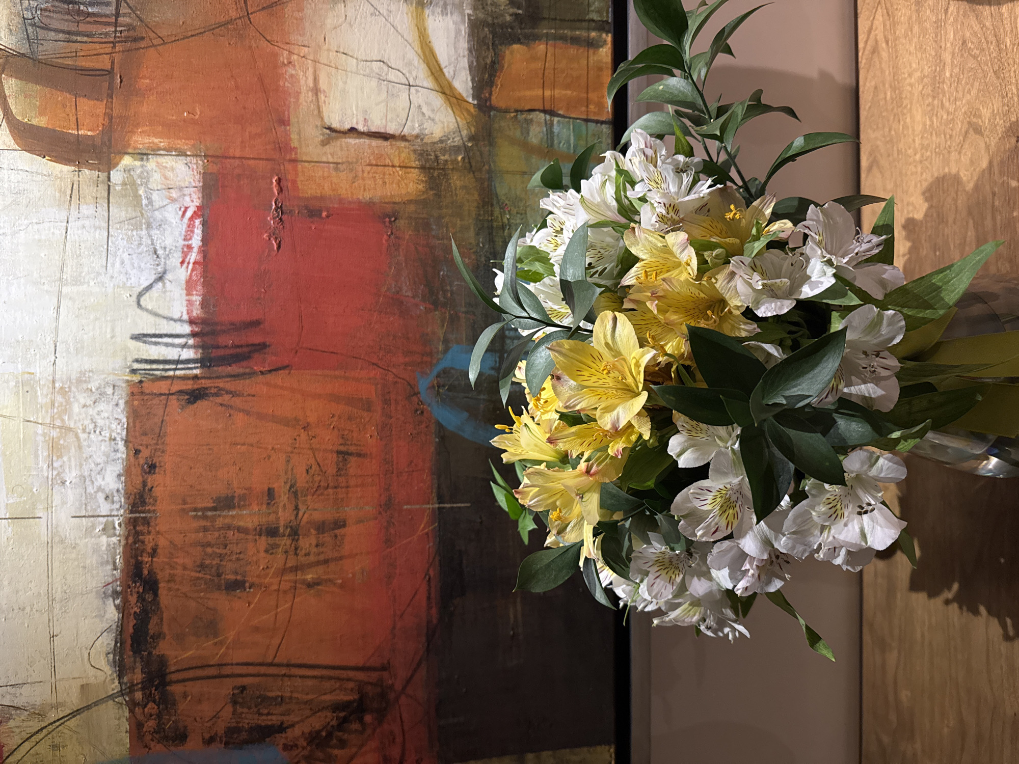 A high-angle shot of a floral arrangement placed in front of an abstract painting. The flowers are a mix of white and yellow alstroemeria, with accompanying green foliage. The painting behind the flowers features a palette of red, brown, white, and black, seemingly with a textured surface. The scene is dimly lit. A brown wooden panel is partially visible on the right edge of the image. The location and time of day are unknown. There are no visible people.