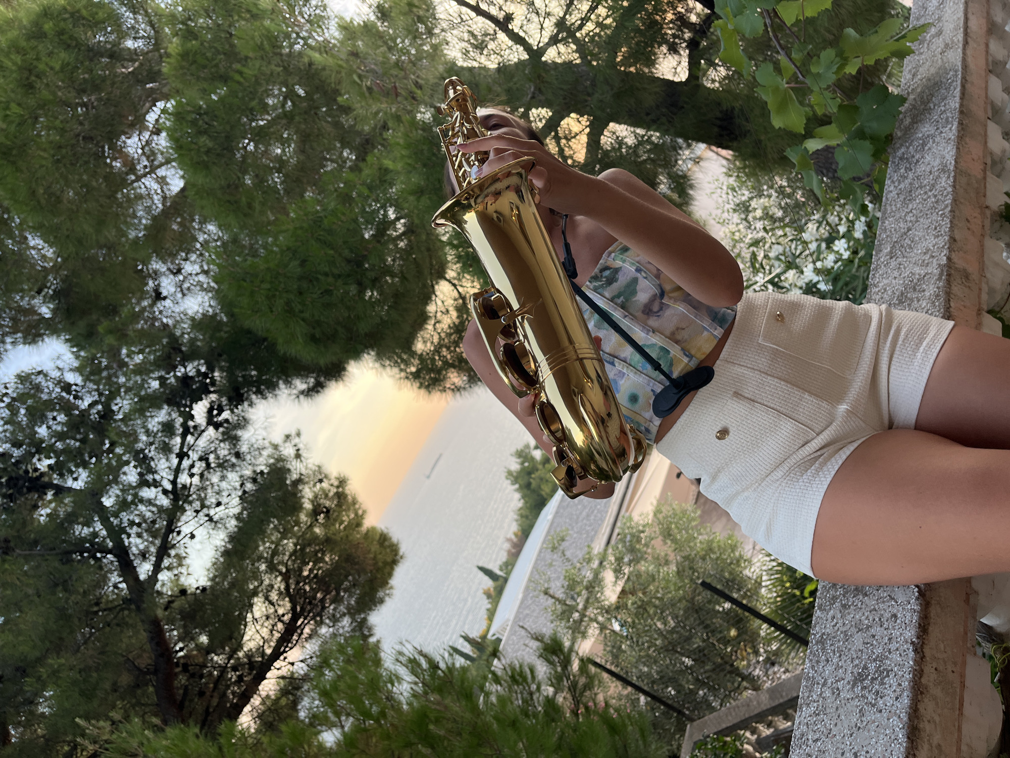 A person, partially obscured by foliage, is holding a golden saxophone in an outdoor setting. They are wearing a patterned top and white textured shorts. The background shows a body of water, likely the Adriatic Sea, with a distant ship and a coastal landscape with buildings and trees. The lighting suggests it is late afternoon or early evening. The location is identified as Šušanj, Montenegro.