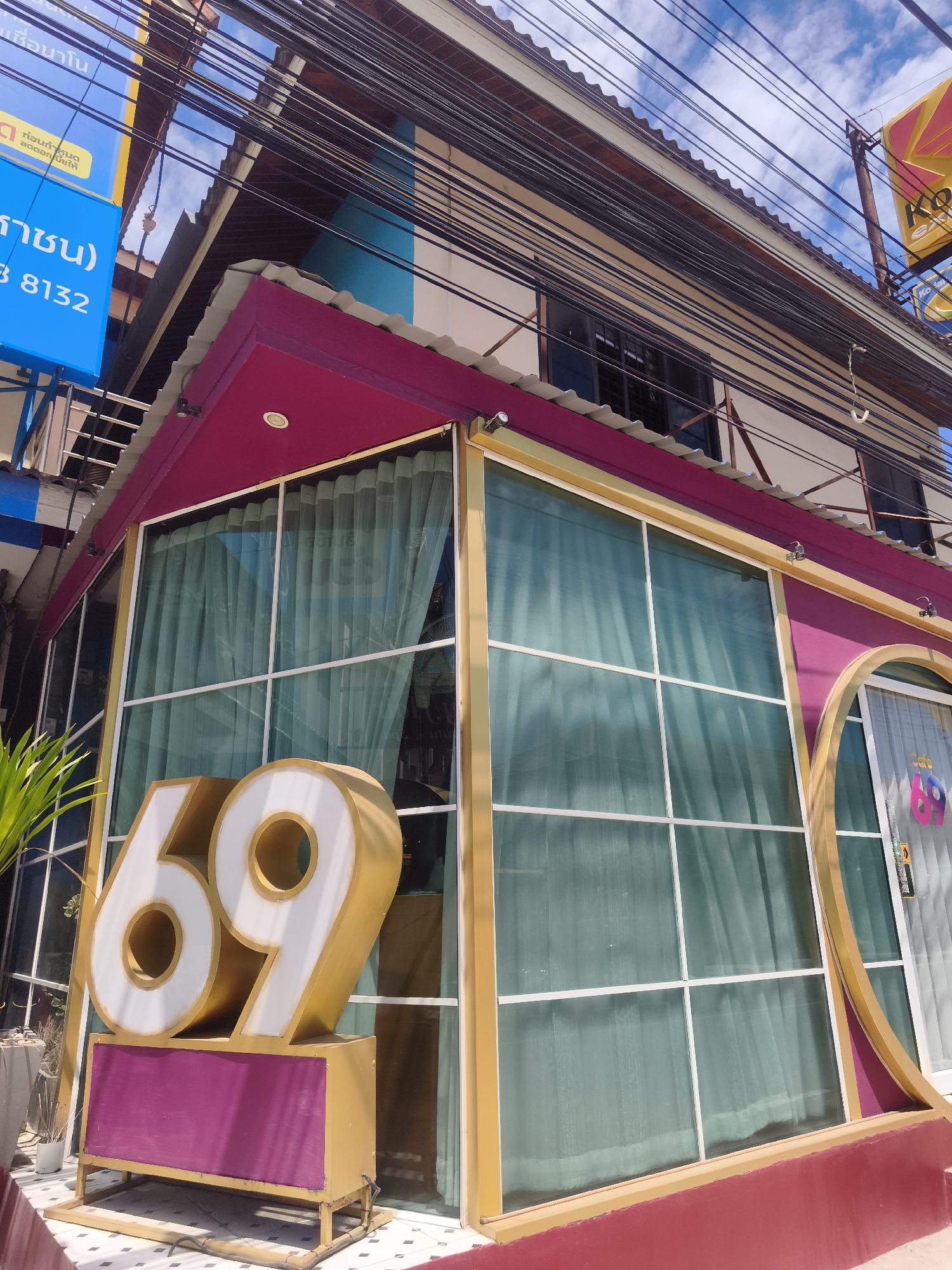 Captured in Baan Bo Phut, Thailand, the image focuses on a building prominently displaying a large, white and gold "69" sign in front. The building’s facade is painted in shades of magenta and gold, featuring large glass windows covered by sheer curtains. Above the windows, electrical wires crisscross against a partly cloudy sky. The building's design includes a rounded doorway and a sign indicating "Cafe 69". The overall scene appears to be an urban setting, with elements such as a blue advertising sign visible in the background, suggesting a commercial or tourist area.