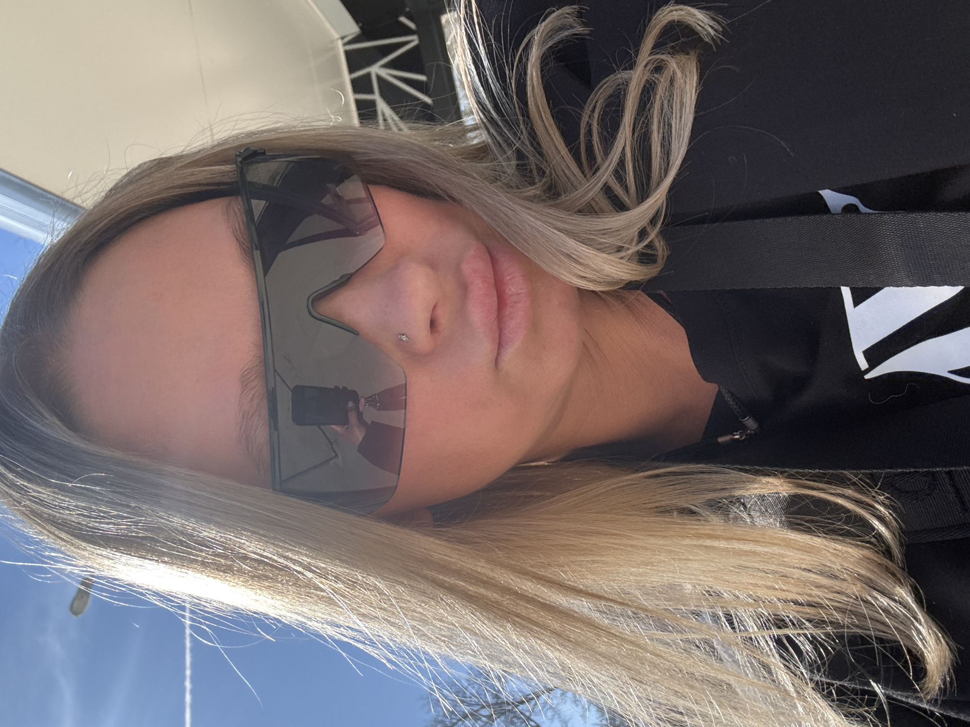A close-up, canted, medium-shot depicts a blonde woman with a nose piercing wearing sunglasses and a black top with a white design. The reflection in the sunglasses shows someone holding a phone. The setting appears to be outdoors under a bright, partly cloudy sky. No elements within the image allow for the location to be identified as Perm, Russia.