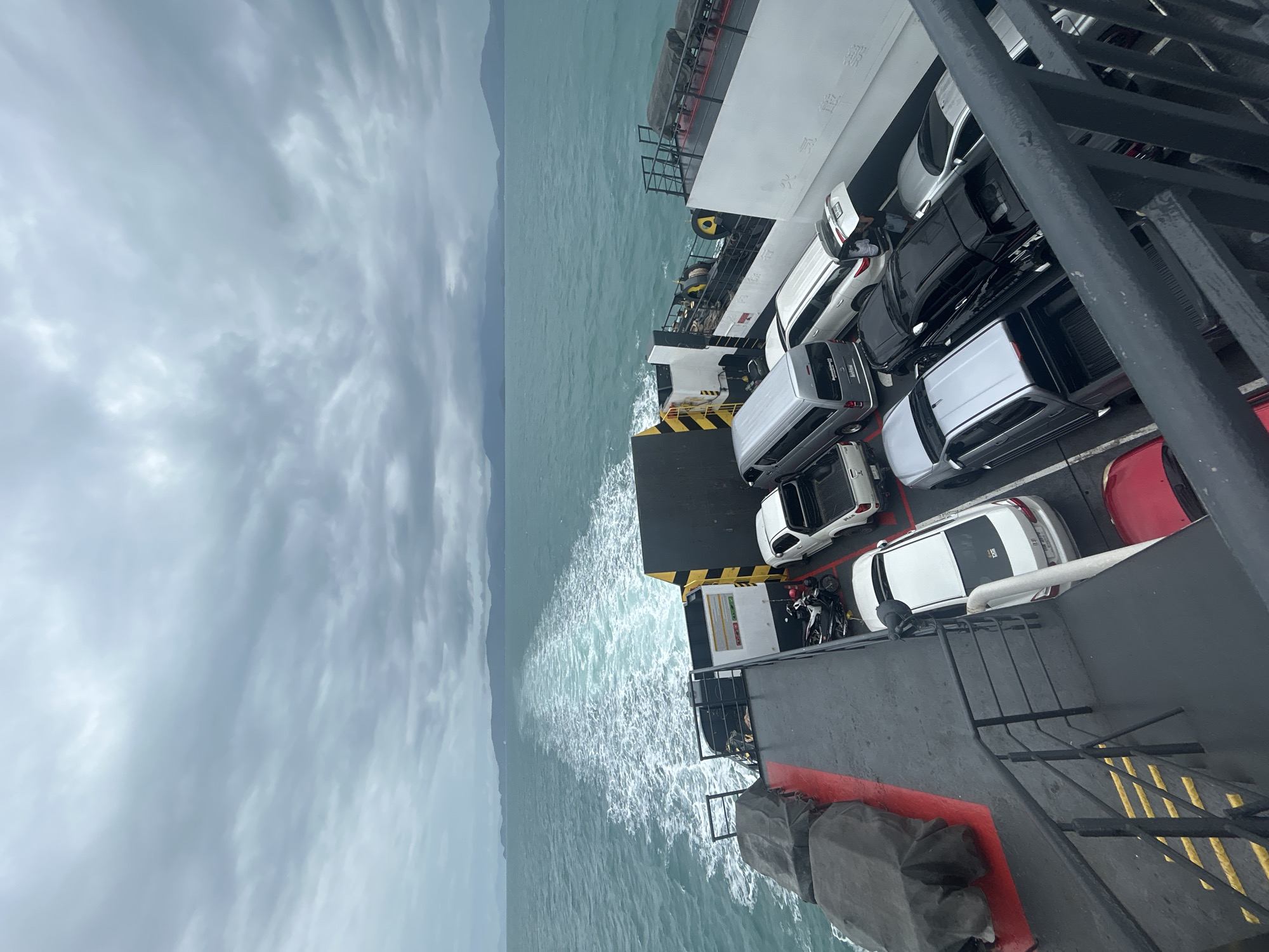 On a cloudy day, a ferry transports several vehicles across a body of water, with a wake trailing behind. The ferry deck, made of dark metal with yellow safety markings, is packed with cars, including white sedans, a white pickup truck, a silver pickup truck, a black SUV, a silver SUV, and a grey minivan. Two motorcycles are parked on the deck. In the distance, a mountainous coastline is visible under a grey, overcast sky. The sea is a muted turquoise, with whitecaps indicating choppy conditions.
