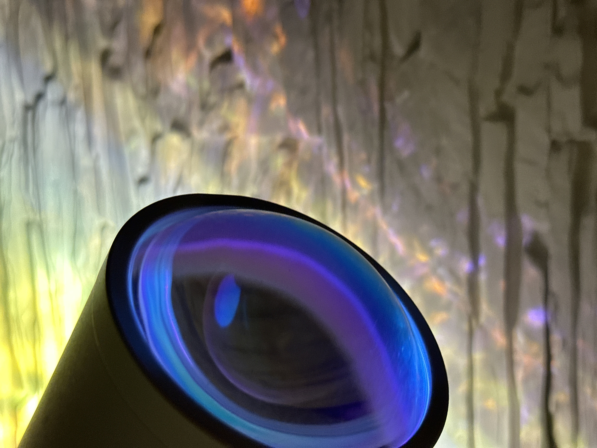 The image displays a close-up, angled view of an optical lens, likely from a camera or similar device, dominating the lower portion of the frame. The lens exhibits a prominent blue and purple anti-reflective coating, creating concentric rings of vibrant color against its dark interior.
The background is heavily out of focus, creating a soft bokeh effect. It features an abstract pattern of vertical ridges or folds, illuminated with a spectrum of colors including soft yellows, oranges, purples, and grays, which appear to shimmer or reflect light. These colorful patterns are indistinct, suggesting either a textured surface like draped fabric or a wall with complex lighting, or perhaps a light projection onto a surface.
No people or discernible activity are present. The image is a still life, focusing on the interplay of light and optics. The lighting appears artificial, highlighting the colorful reflections and coatings. There are no clear cues for time of day, weather, or a specific outdoor location; the setting seems to be indoors or in a controlled environment. No visible text is present.