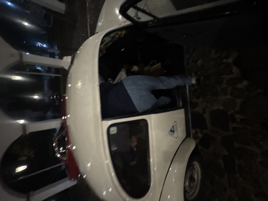 The scene depicts a nighttime view within an arched, colonnaded area with cobblestone paving, characteristic of colonial architecture in Taxco, Mexico. A white classic Volkswagen Beetle is positioned with its driver's side door ajar. A person's lower body, clad in blue jeans and light-colored footwear, is observed in the process of entering or exiting the driver's seat. A faint outline of another person's hand is visible through the rear driver's side window. In the background, under illuminated white arches, a red classic car is parked. Artificial lighting provides illumination against the dark environment.