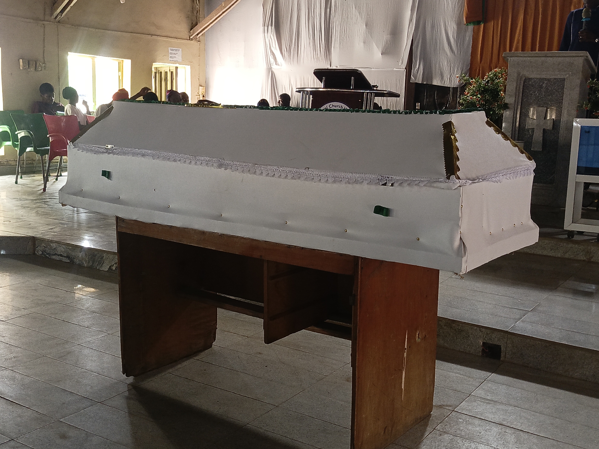 The image depicts an indoor scene, likely a funeral service or wake, set within a church or a large hall. In the foreground and central to the image is a white, fabric-draped coffin, resting on a simple dark wooden stand. The coffin is decorated with gold trim, white lace detailing, and small green accents on its sides.
In the background, several individuals are present. On the left side, two people are clearly visible, seated on green and red plastic chairs, with another partially obscured figure behind them. Heads of other attendees are visible in the mid-background, positioned behind the coffin. To the far right, a person wearing a dark blue top is partially seen behind a grey stone stand that features a carved cross, and they appear to be holding a microphone, suggesting they are speaking or leading the service.
A dark wooden pulpit or lectern is situated in the mid-background, with the word "Church" faintly visible on its side. The interior space is lit by natural daylight streaming through windows in the background. White and orange/brown fabrics are draped as decoration, and parts of the ceiling show exposed wooden beams. The floor is tiled. The overall setting and the presence of the coffin indicate a solemn event.