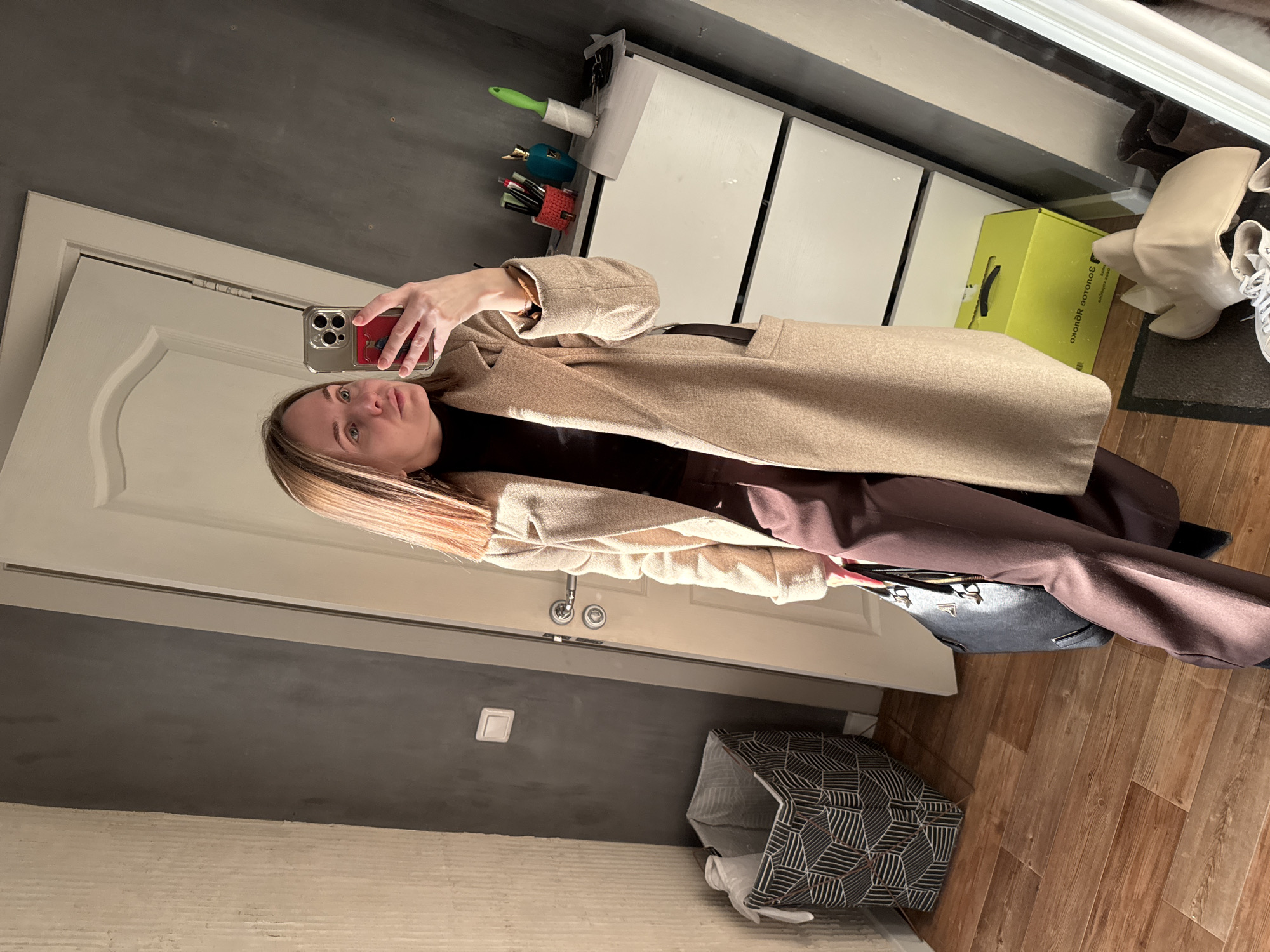 A woman is taking a selfie in front of a mirror inside an apartment in Perm, Russia. She is wearing a brown turtleneck, brown pants, and a tan coat with a black belt. Her hair is blonde. She is holding a cell phone with a red phone case. The mirror reflects a white storage unit with drawers, a green and yellow box, and a pair of white boots and white sneakers. To the left of the woman is a closed, white door and gray wall. At the bottom right of the photo, there is a patterned gray and white laundry hamper and wooden floors. A light switch is on the left gray wall.