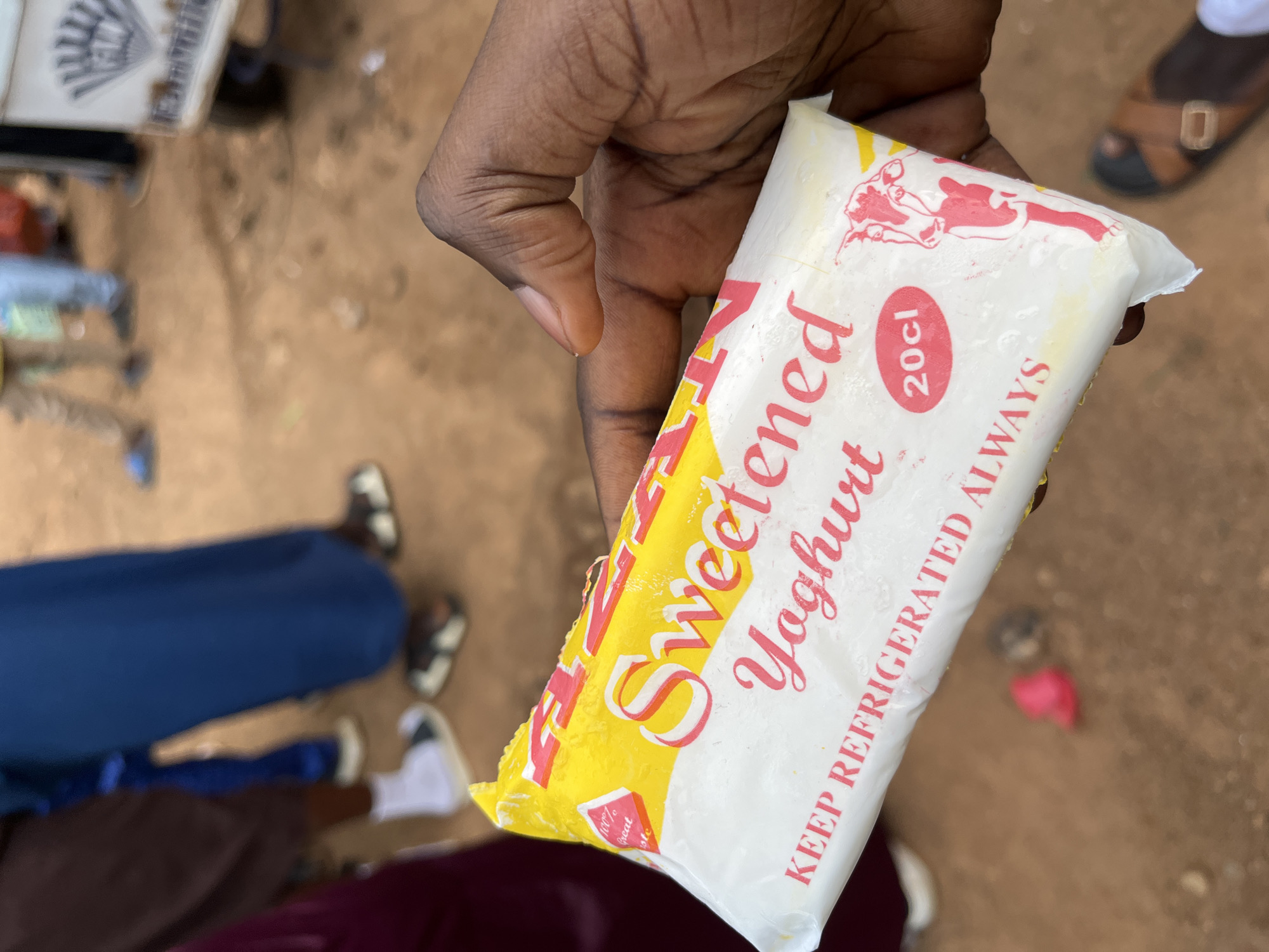In the city of Jos, Nigeria, a person's dark-skinned hand holds a white and yellow packaged yogurt bar. The packaging features pink lettering that reads "Sweetened Yoghurt" and "KEEP REFRIGERATED ALWAYS". A red circle with "20cl" inside is visible, along with a pink and white drawing of a cow. In the blurred background, several people are standing on a dirt ground. One person is wearing a blue garment, another is wearing a dark brown skirt and white socks with sandals, and another is wearing a darker burgundy skirt. A white cart with a fan-like emblem is also visible in the background. The ground appears to be a reddish-brown earth.