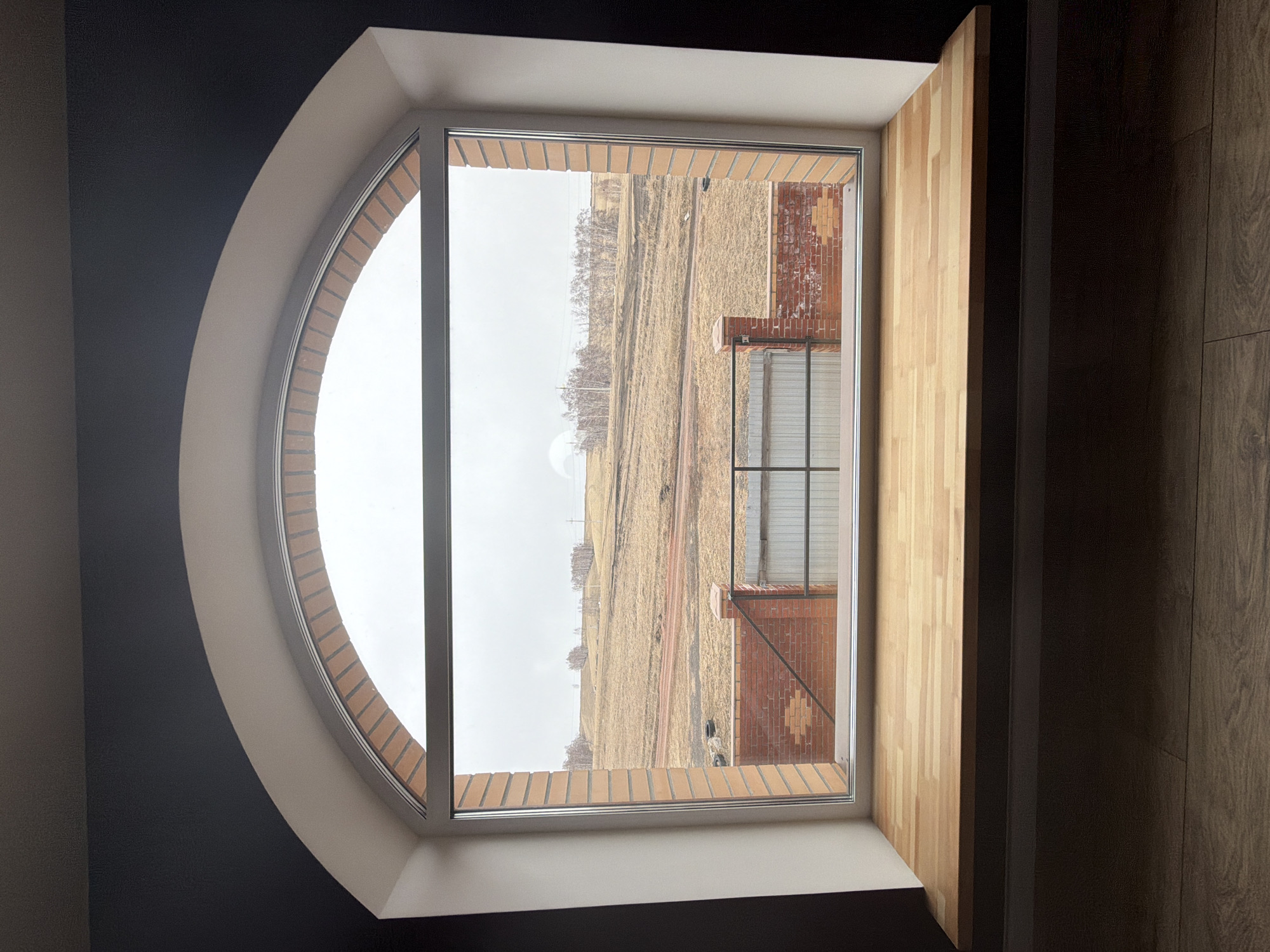 The image shows an interior view through a unique arched window, likely a feature of the architecture. The window has a semi-circular top and a rectangular bottom portion. The interior surrounding the window consists of dark walls on the left and wooden panels on the right.
The view through the window is of an outdoor rural landscape. The immediate foreground outside the window includes a brick structure. Beyond that, the landscape features dry, grassy fields and some sparse trees. There also seems to be a red dirt or gravel path winding through the fields. The sky is overcast and appears gray or white, suggesting cloudy weather.
The location in Krasnoyarsk, Russia is not evident from the content of the image.