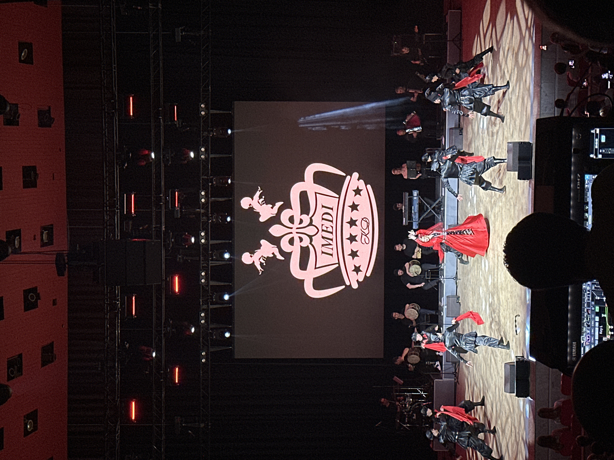 This is a photograph of a stage performance at the Imereti Concert Hall in Sochi, Russia. The stage features a large screen displaying a logo with the text "IMEDI" and a crown. In front of the screen, a group of performers are dancing, dressed in black costumes with red sashes. To the left, a band is playing musical instruments, including drums and an accordion. In the foreground, an audience is visible, seated in a dimly lit hall. The stage is lit by spotlights, creating dramatic shadows. The overall atmosphere appears to be that of a cultural performance or concert.