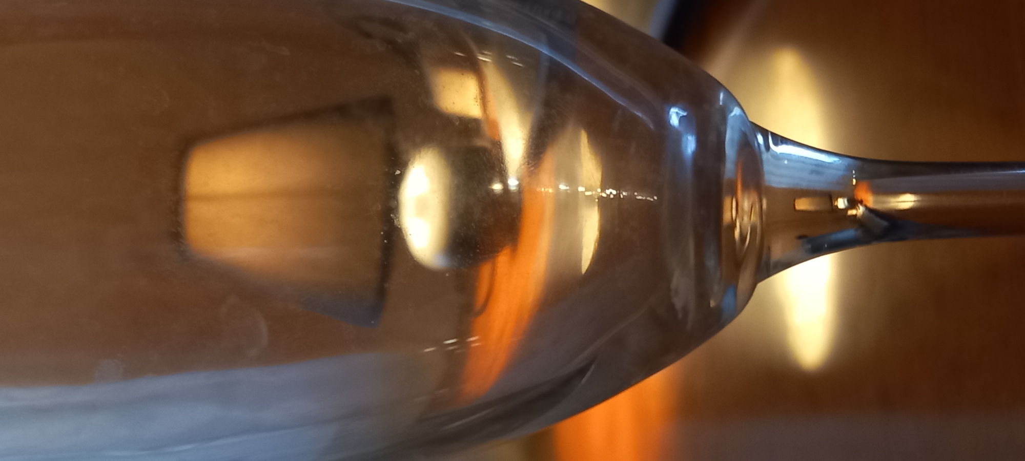 The image displays a close-up, horizontally oriented shot of a clear glass, likely a wine glass or champagne flute, resting on a warm-toned surface. The primary focus is on the intricate interplay of light, reflections, and refractions through the glass.
The background is a soft, uniformly lit, warm brown or ochre hue, suggesting a wooden surface or wall, rendered with a shallow depth of field. This surface warmly reflects the ambient light.
Within the transparent body of the glass, several bright artificial light sources are reflected and distorted. A prominent rectangular or square light source is visible on the left, appearing diffused and elongated by the glass. Towards the center, a distinct, bright, circular reflection stands out, creating a glowing effect. Numerous smaller, scattered highlights shimmer across the curved surface of the glass, indicating multiple points of illumination. The stem of the glass extends towards the right, also catching and reflecting light.
Subtle details on the glass surface include faint smudges and small particles of dust, visible upon closer inspection. The overall lighting scheme is warm and artificial, characteristic of an indoor setting, possibly during the evening or in a dimly lit room. No specific activity or event is taking place; the image is a still life composition exploring light and material. There is no visible text within the frame. The location context for this media is Kryvyi Rih, Ukraine.