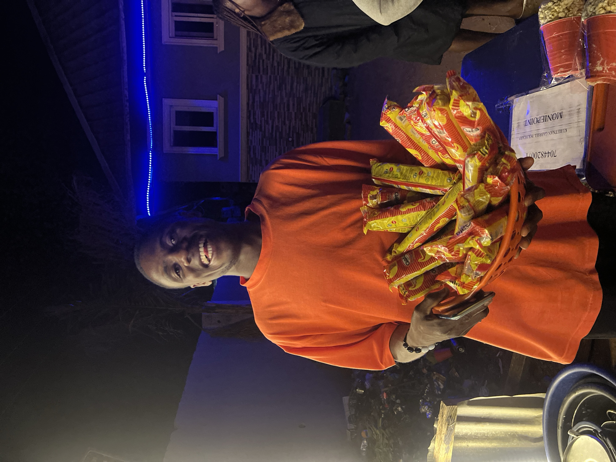 In Durumi, Nigeria, a young man wearing an orange t-shirt stands holding a basket filled with packaged noodles. He is smiling at the camera. Behind him, a building is illuminated by blue LED lights. To his right, another person is partially visible, wearing a black jacket with a fur collar. The scene appears to be an outdoor market stall at night.