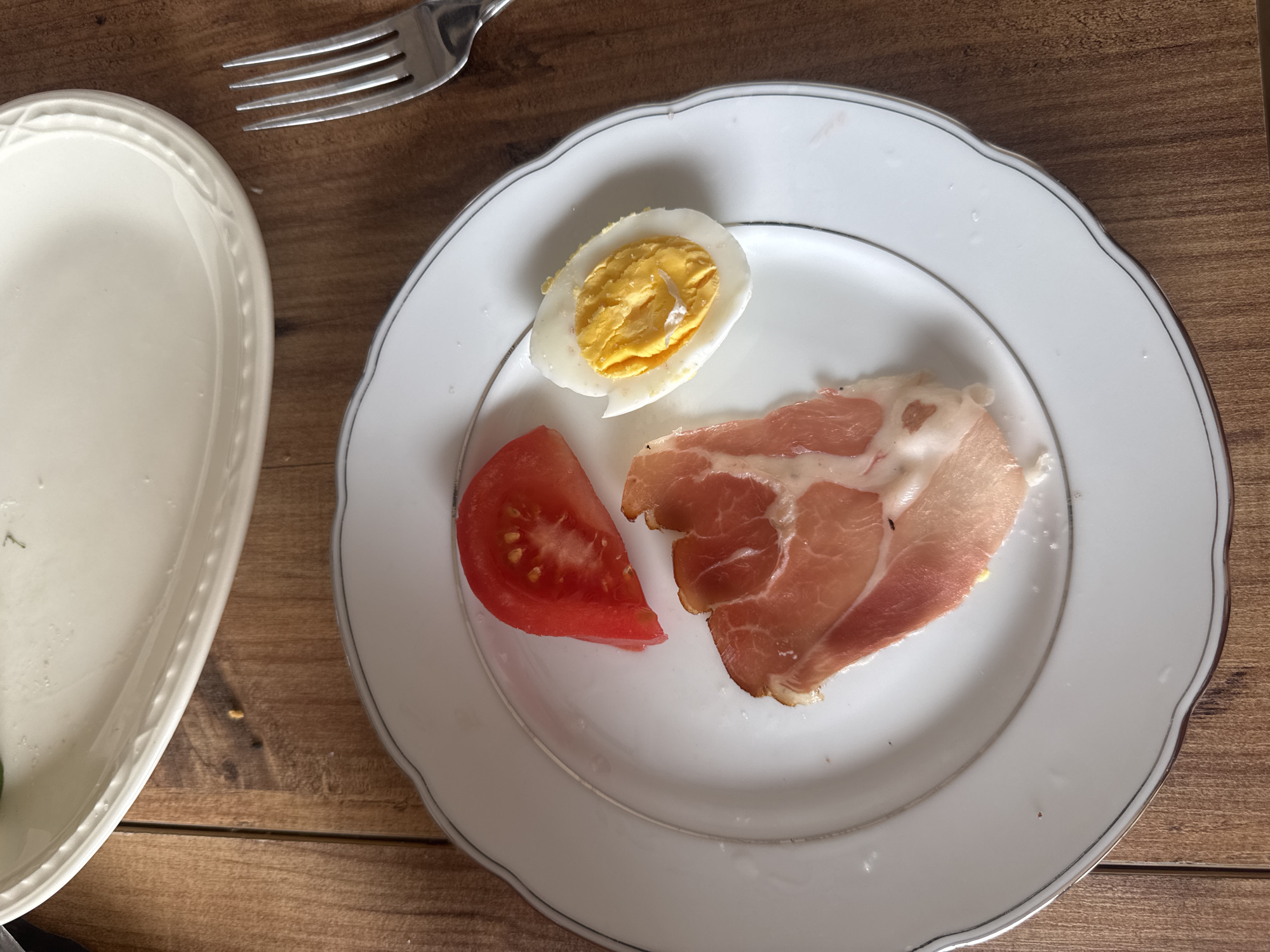 A top-down shot shows a white plate with a silver rim, resting on a wooden surface. The plate contains a half of a hard-boiled egg, a slice of tomato, and a piece of prosciutto. To the left is a portion of a second white plate, and at the top left, the tines of a fork are visible. The scene appears to be a breakfast or light meal setting in an indoor environment. Location: Unknown, Unknown.
