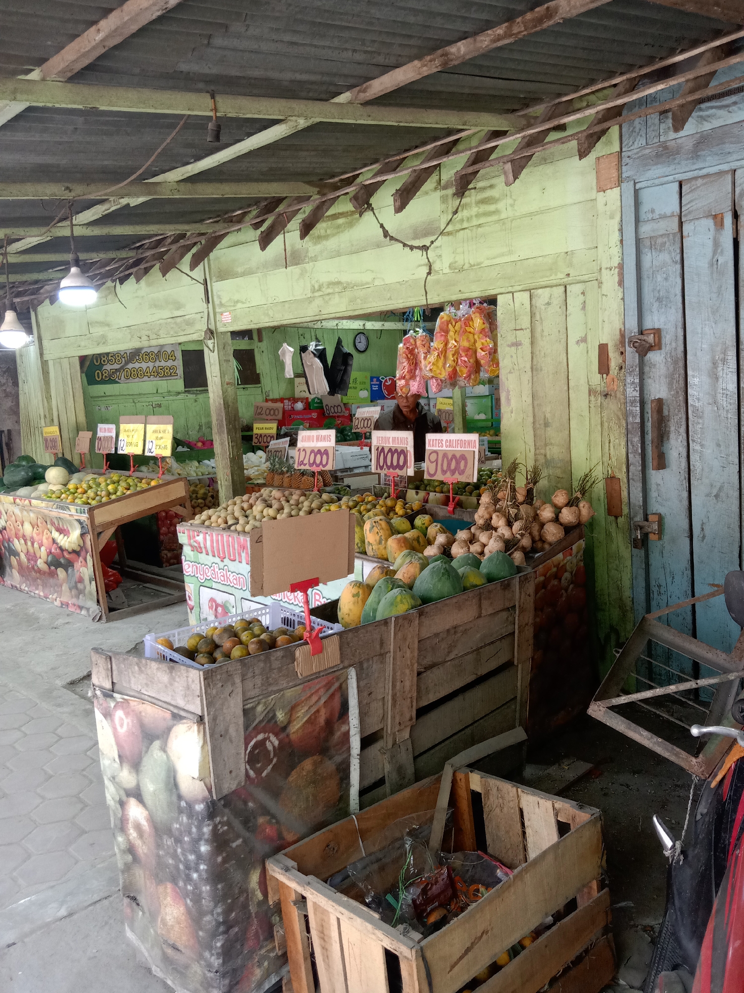 This is a medium-shot, eye-level, outdoor, natural-light photograph of a fruit stand in Sugihwaras, Indonesia. The stand is constructed from weathered wood and appears to be made of several different sections. The front of the stand is lined with various fruits and vegetables, including oranges, papayas, and what appear to be sweet potatoes. The fruits are displayed in wooden crates and bins. Signs with prices in Indonesian Rupiah are posted above some of the fruit displays. A man is partially visible behind the counter, wearing a dark shirt and a hat. In the background, there are more fruits and vegetables, as well as some hanging bags of what look like snacks. A large wooden door with blue-painted planks is on the right side of the frame. A metal rack is leaning against the door, and a motorcycle is partially visible on the far right. The overall impression is of a bustling, local market.