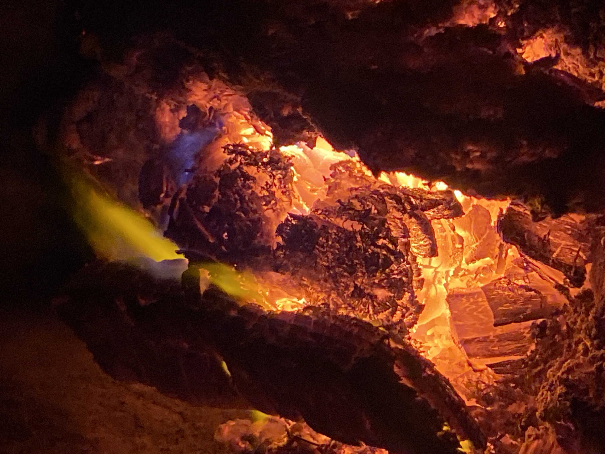 The image depicts a close-up view of a campfire with brightly burning embers and flames. The fire is contained within what appears to be a natural setting, with dark, textured logs forming the structure of the fire pit. Intense orange and yellow hues dominate the scene, indicating high heat. Hints of blue light are visible within the flames, suggesting the presence of certain chemical elements or cooler combustion zones. The background is largely obscured by darkness, emphasizing the illuminated core of the fire. There are no people or animals visible in the image, nor are there any man-made structures or indications of an urban environment. The scene is entirely focused on the natural phenomenon of fire. No information regarding the city of Nyagan, Russia, can be derived from this image.
